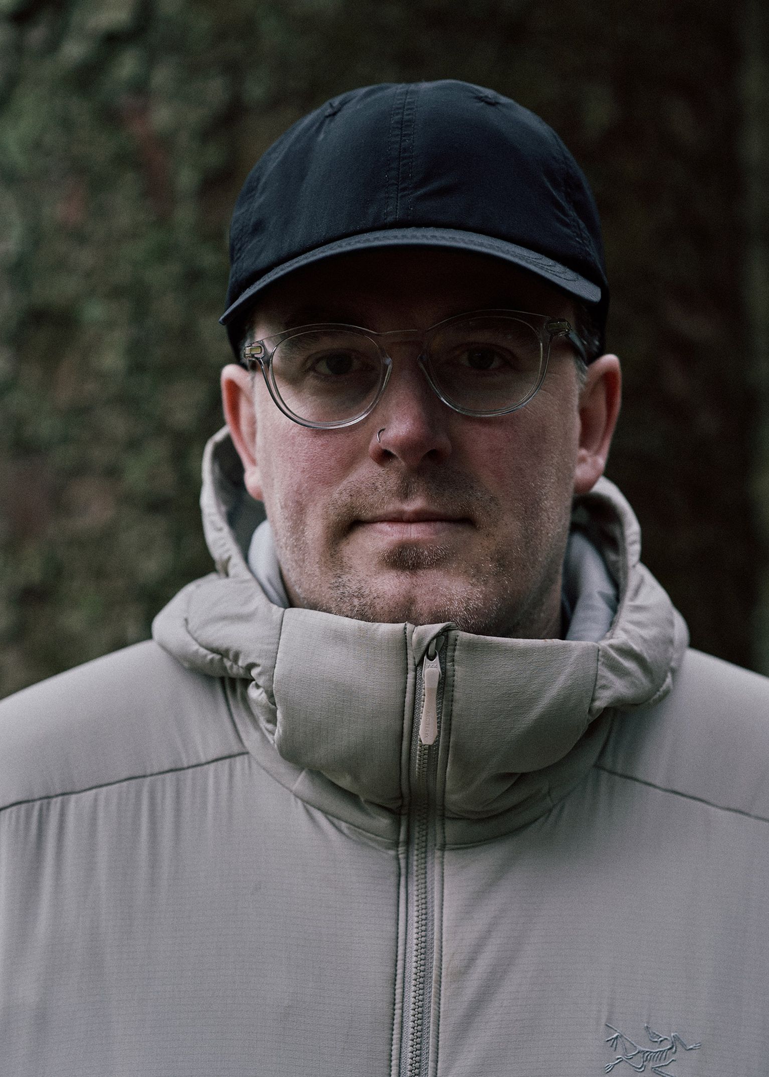A person wearing a black cap, glasses, and a light grey puffer jacket stands outdoors in front of a large tree trunk.