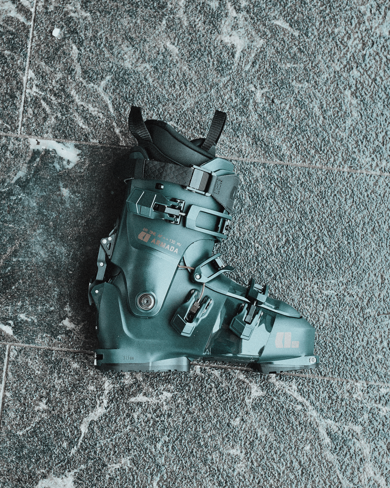 Green ski boot with black buckles and orange accents on a textured grey floor.