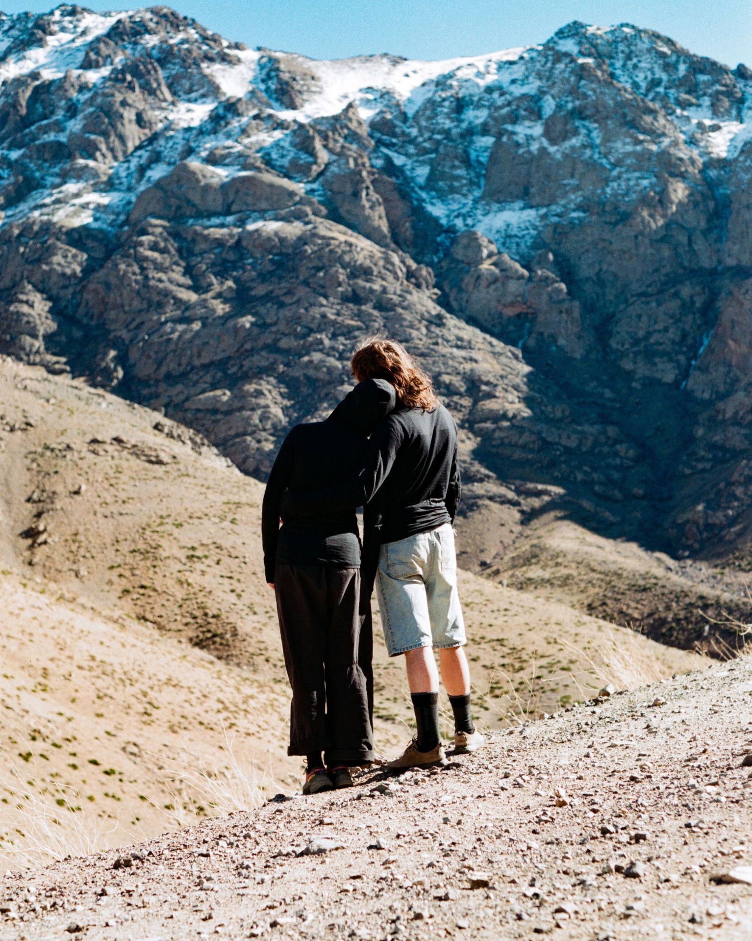 Two people stand on a rocky mountain path, embracing, with a snow-dusted peak in the background under a clear blue sky.