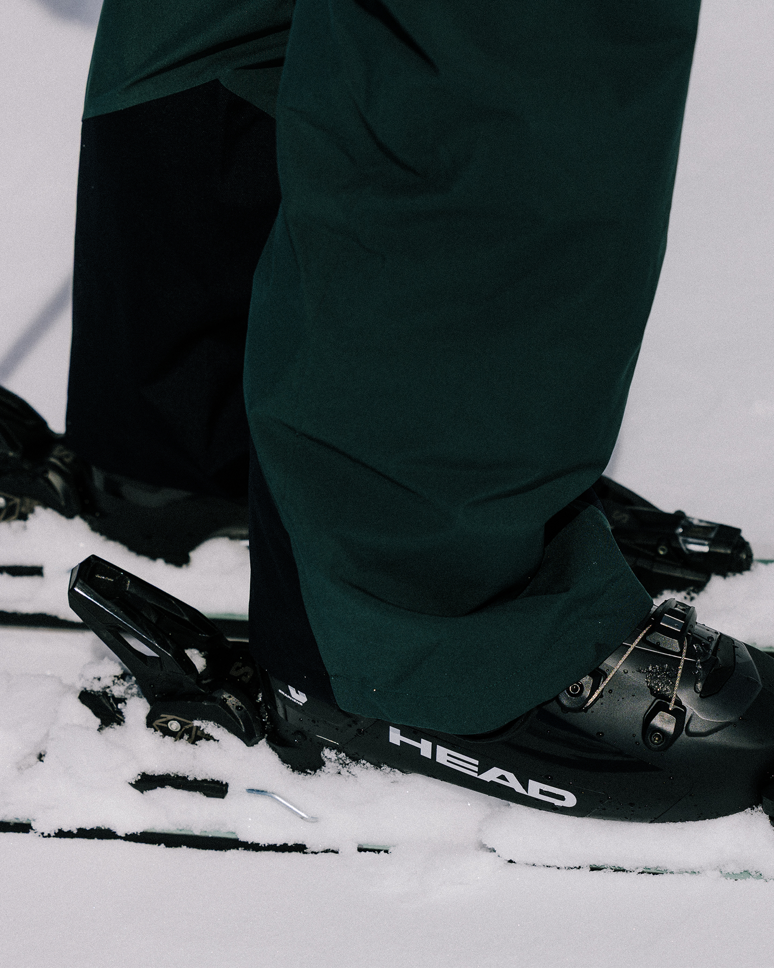 Close-up of a skier wearing black ski boots and dark green trousers, standing on skis with bindings in the snow.