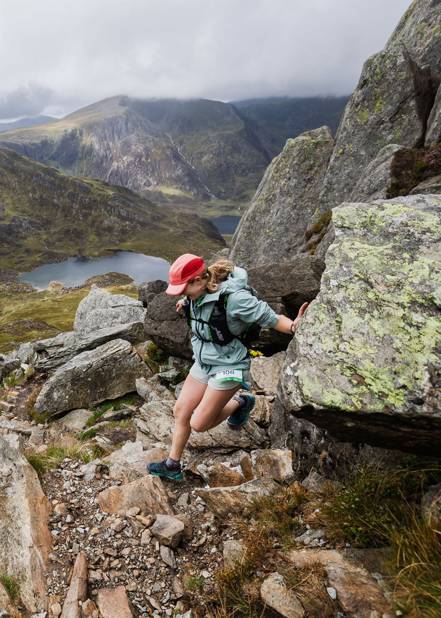 Person climbing rocky mountain trail with scenic view of rugged peaks and a lake in the background, wearing outdoor sports gear.