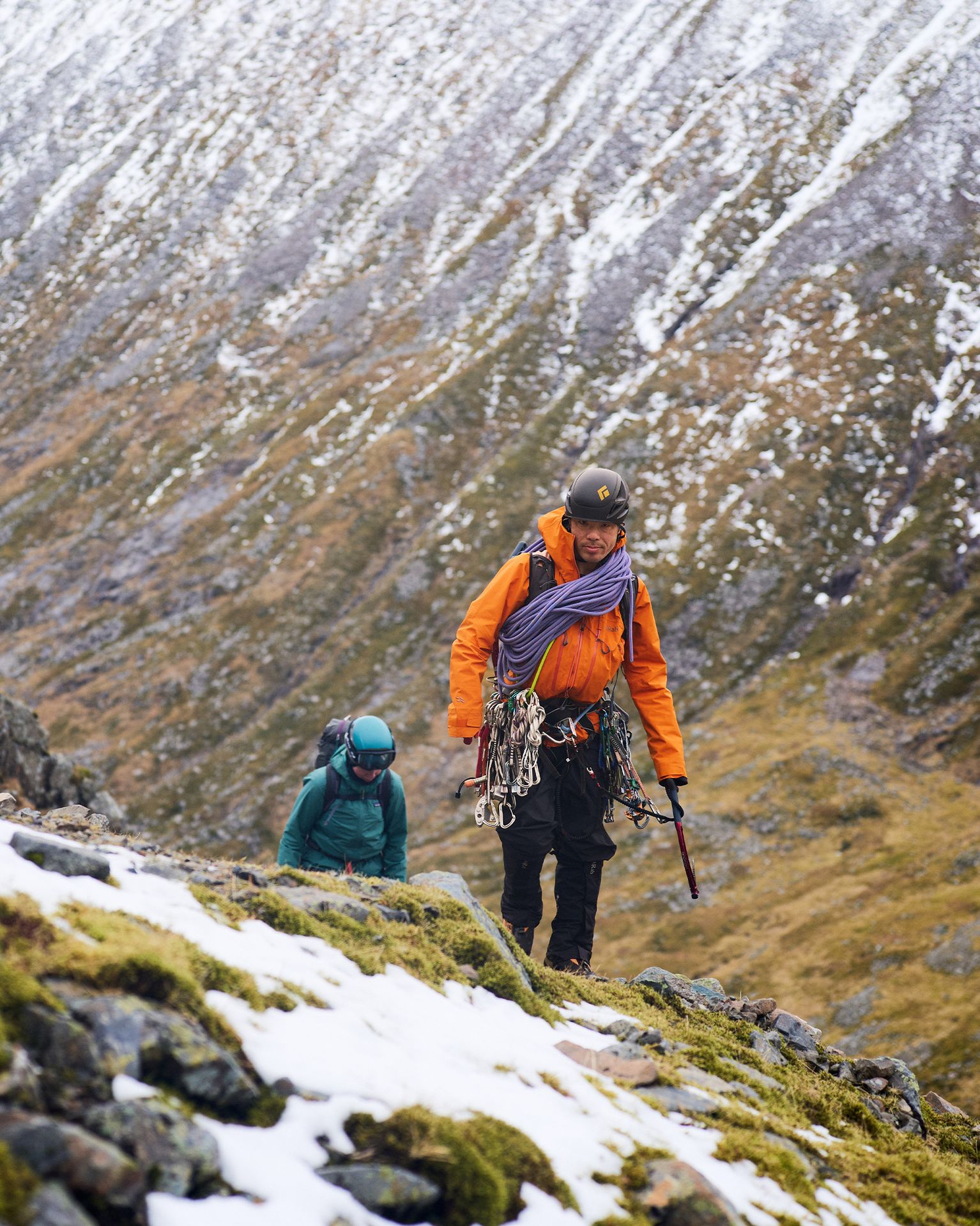 Two climbers in winter gear ascend a snowy, rocky mountain slope, carrying ropes and equipment, with rugged terrain in the background.