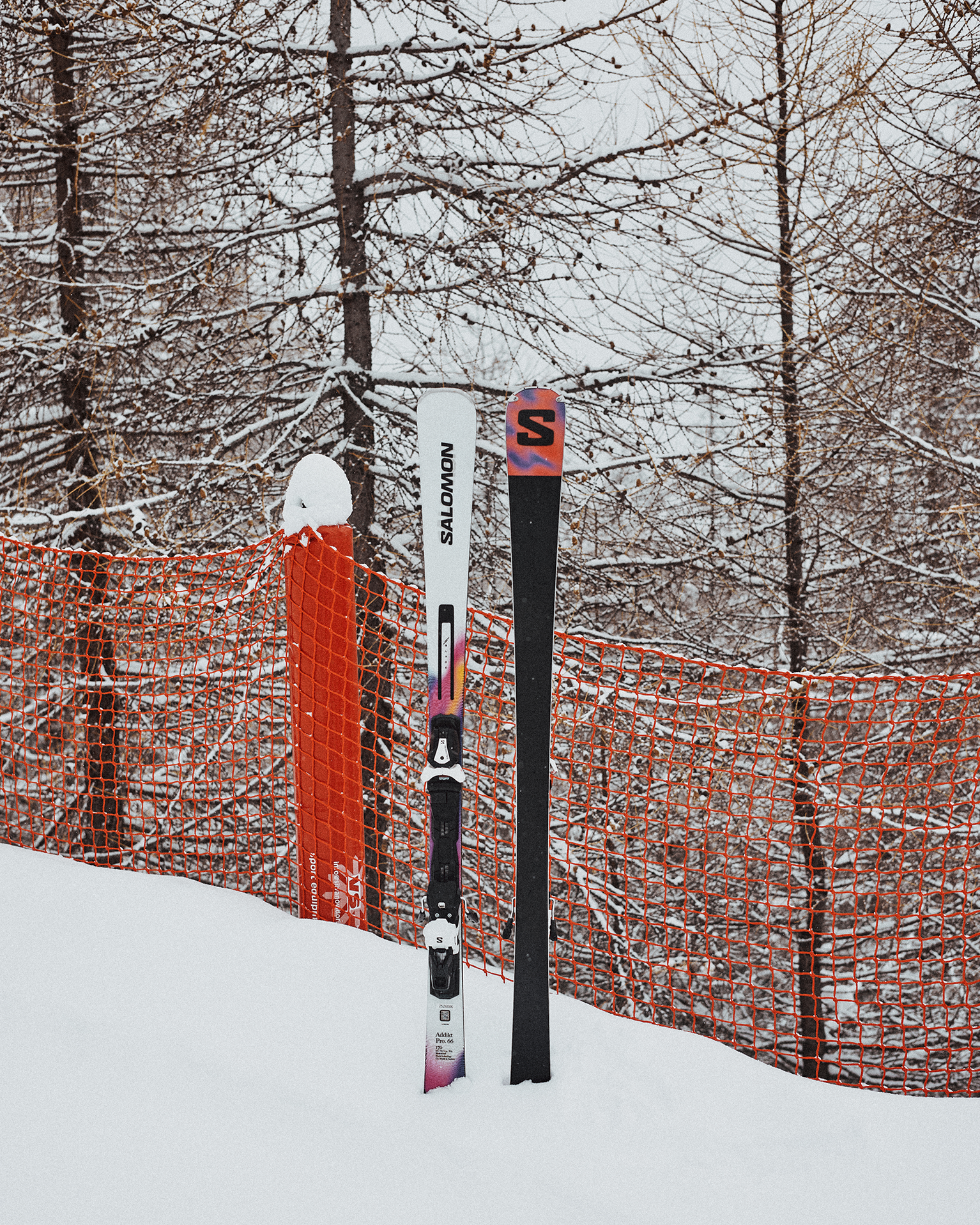 Skis leaning against an orange safety net on a snowy slope, surrounded by leafless trees.