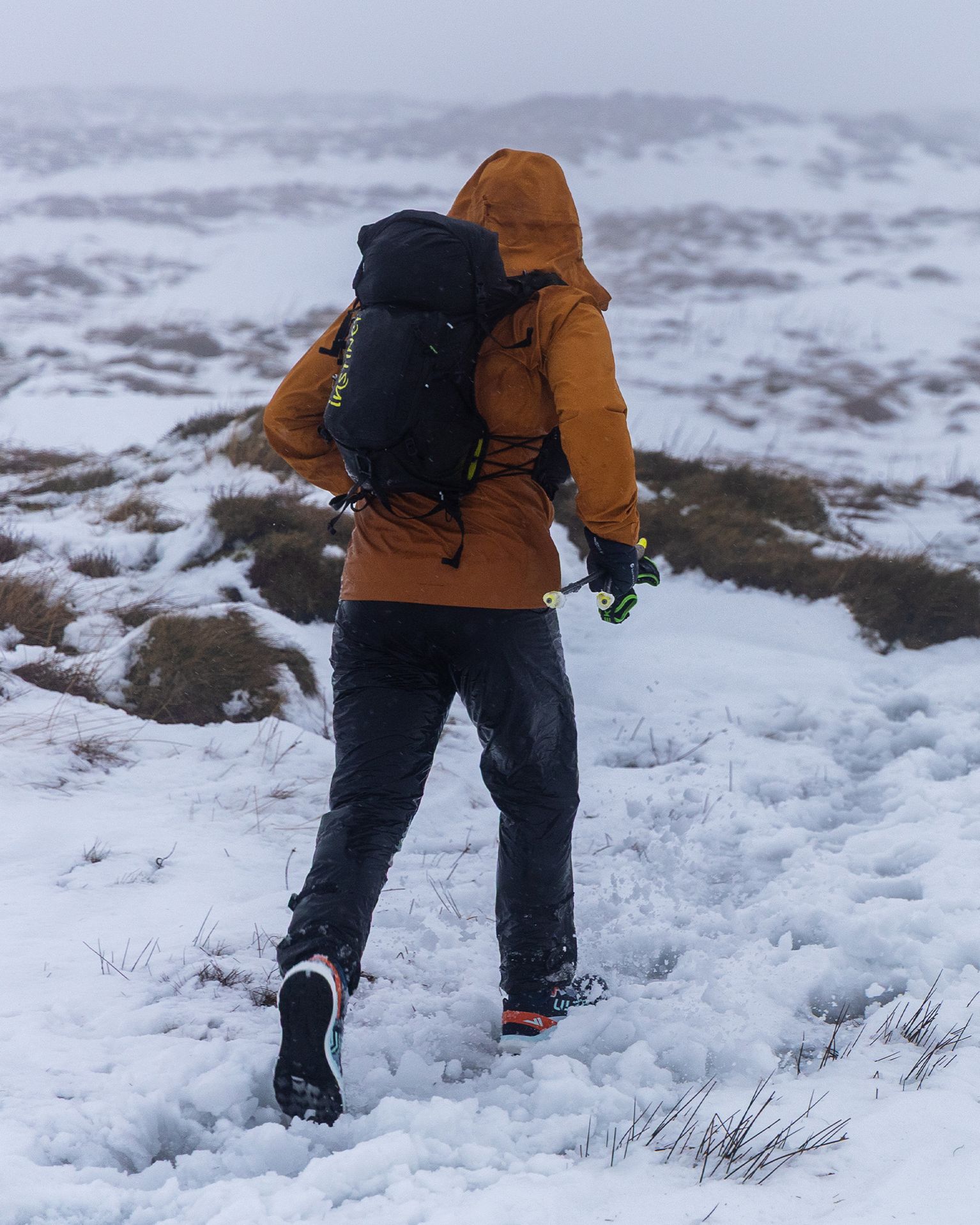 Person in orange jacket and black trousers running through snowy, mountainous terrain, wearing a backpack.