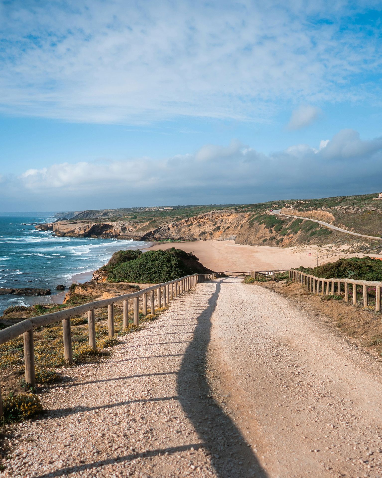 A sandy path leads to a secluded beach with rugged cliffs and ocean waves under a bright blue sky.