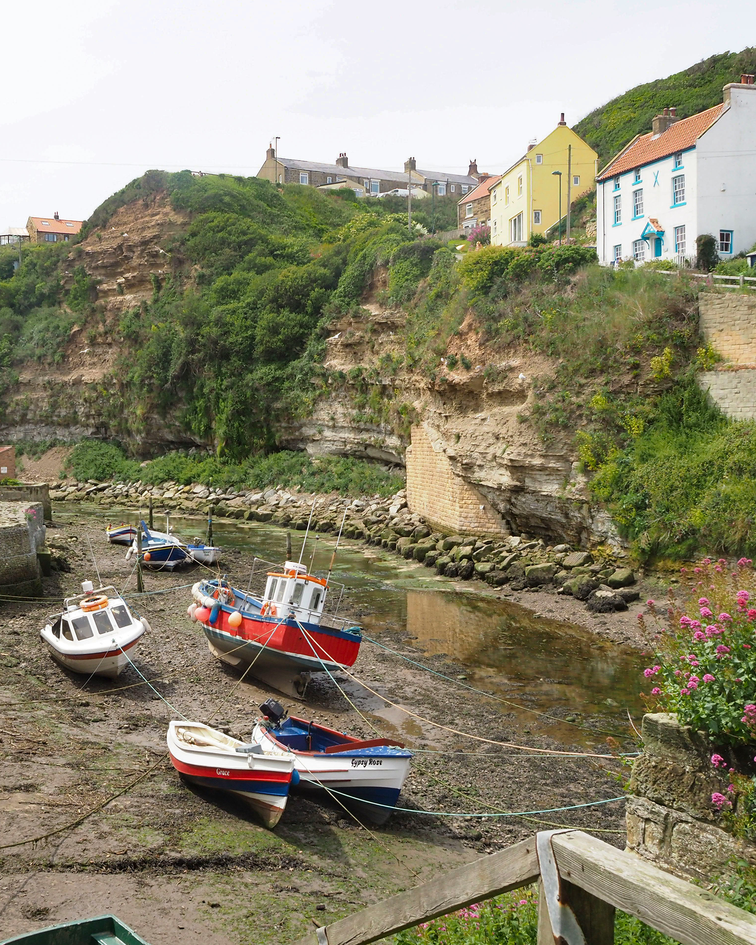 Boats resting on a rocky shore in front of colourful hillside cottages, surrounded by lush greenery and a small winding stream.