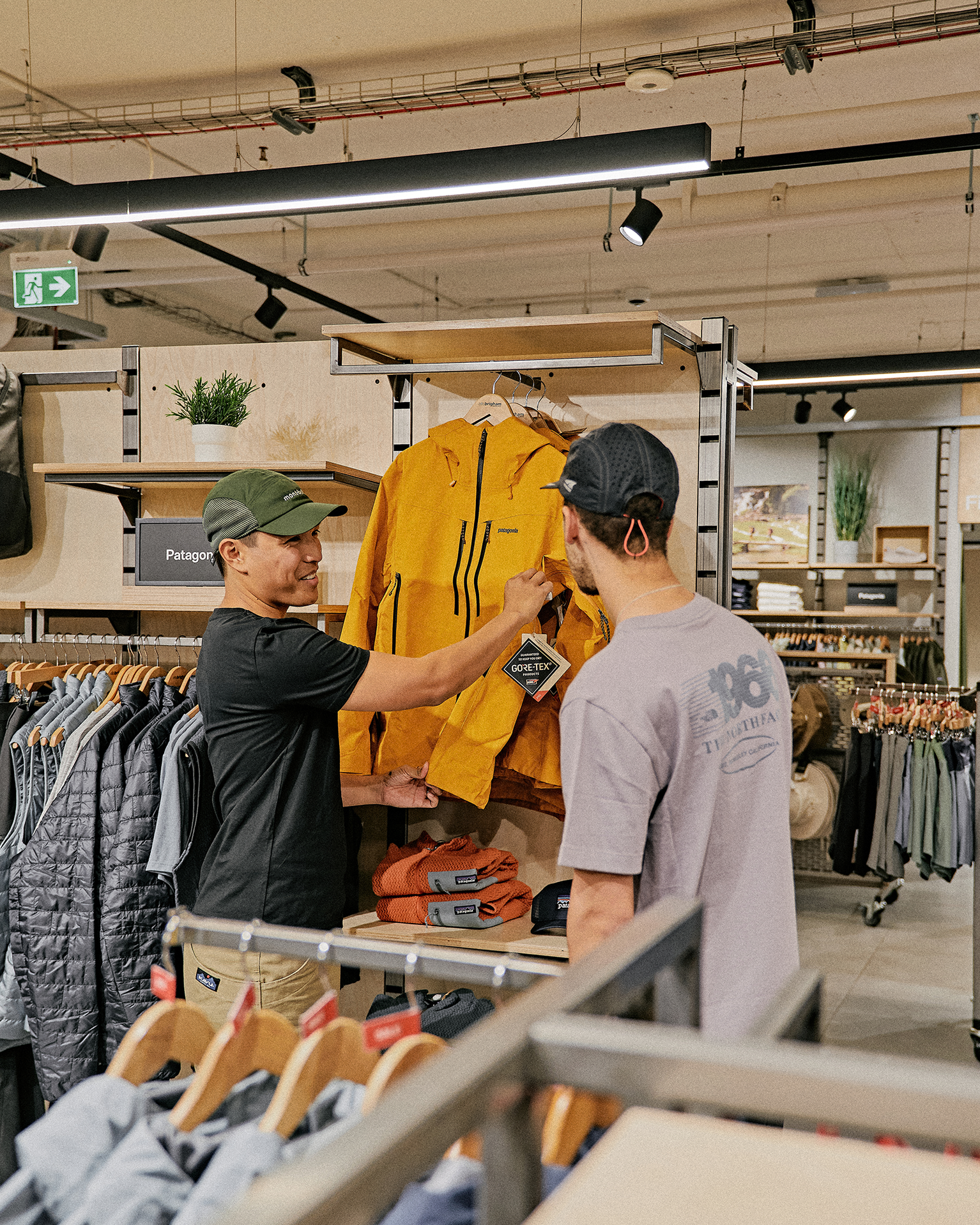 Two people in a store discussing a yellow mountain jacket. Various outdoor clothing items are displayed around them.