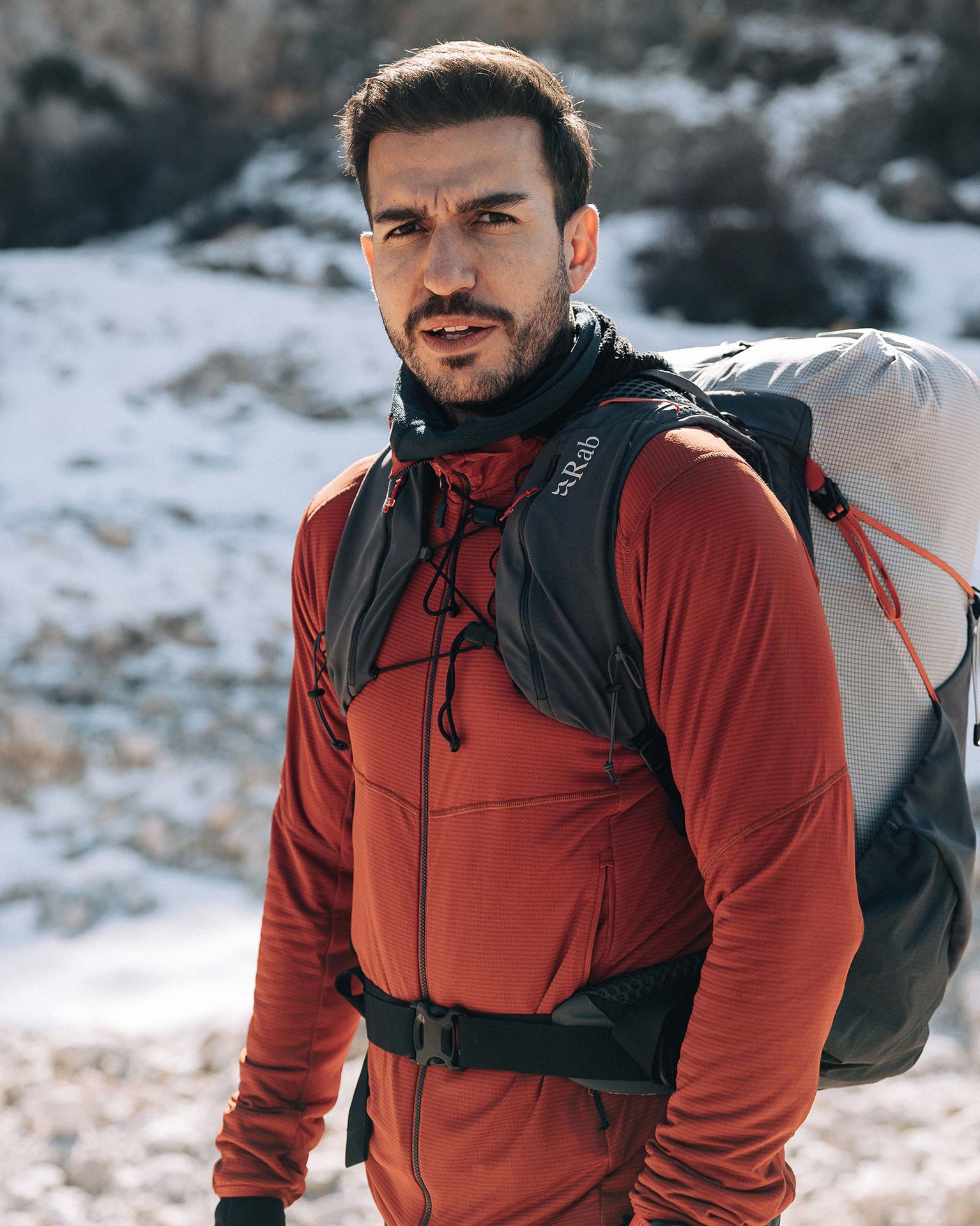 Man wearing a red jacket and a backpack, standing outdoors in a snowy mountain landscape.