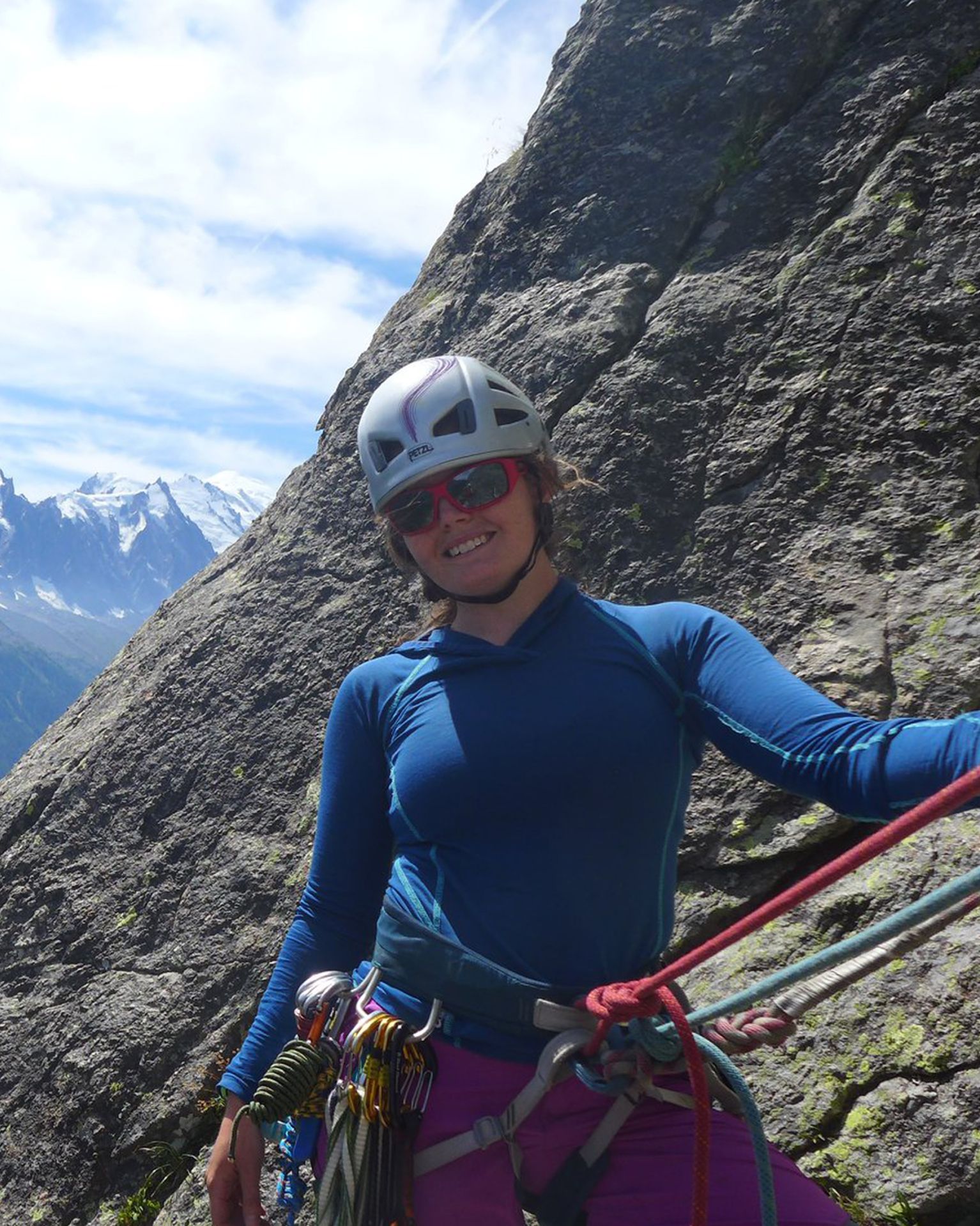 Woman in blue climbing gear smiles while rock climbing against a rugged mountain backdrop.