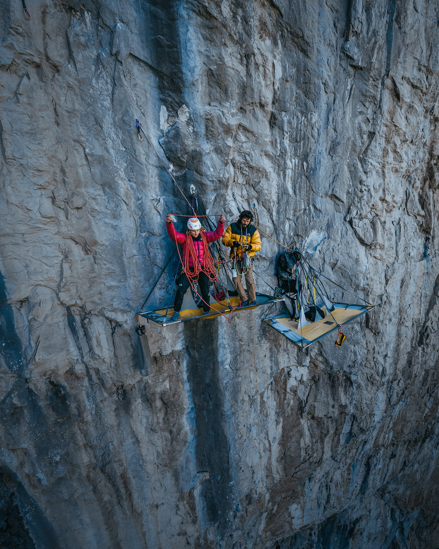 Two climbers on a portaledge scaling a sheer rock face, surrounded by ropes and gear, exemplifying adventure and expertise in challenging terrain.
