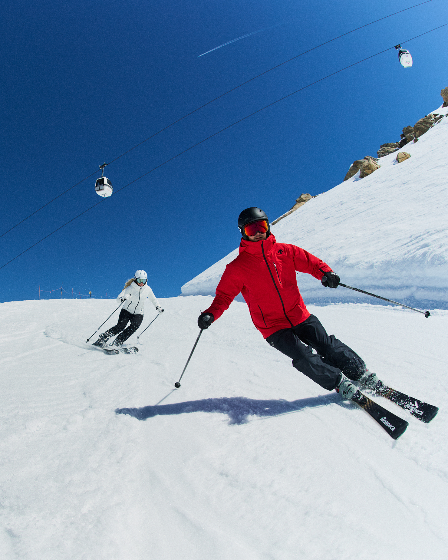 Two skiers in red and white gear glide downhill on a sunny day, with cable cars above and snowy slopes in the background.