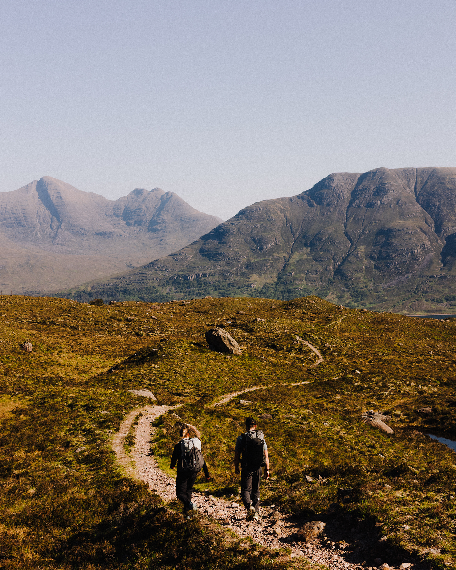 Two hikers on a rocky trail in a vast, sunny mountain landscape with clear skies and rugged peaks in the background.
