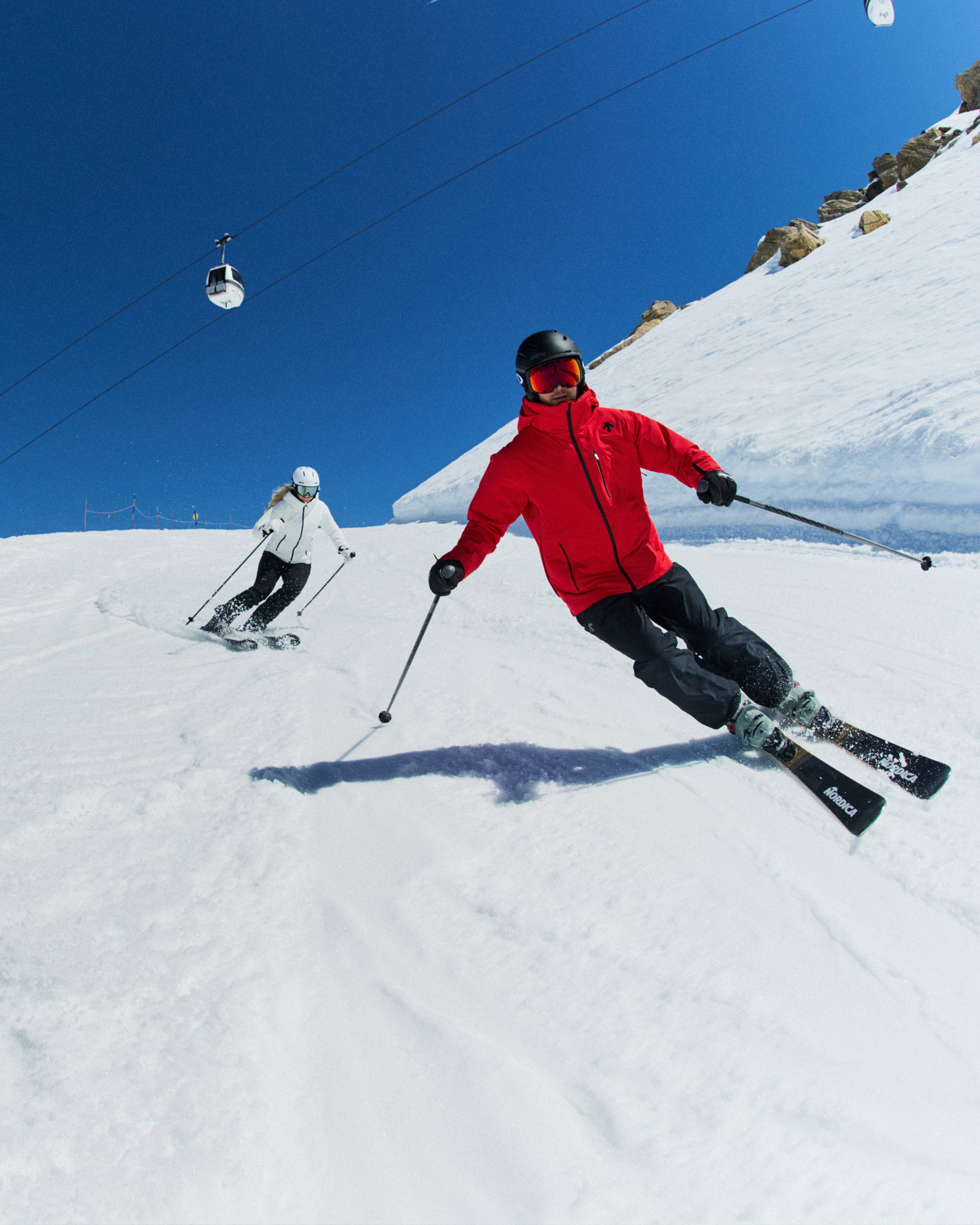 Two skiers in red and white Descente ski gear glide down a snowy slope under a clear blue sky, with a cable car visible above.