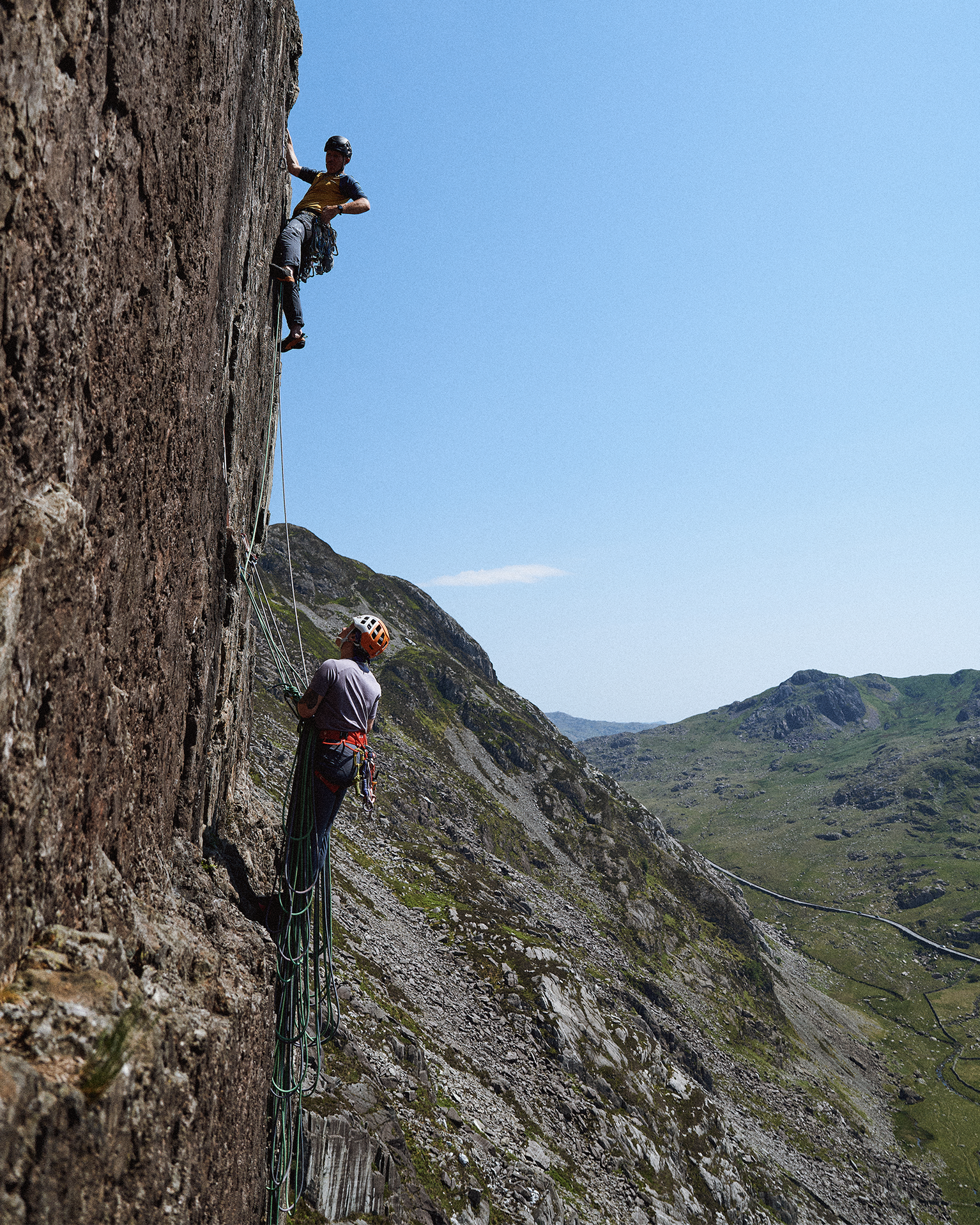 Two rock climbers scaling a steep cliff face in a mountainous landscape under a clear blue sky.