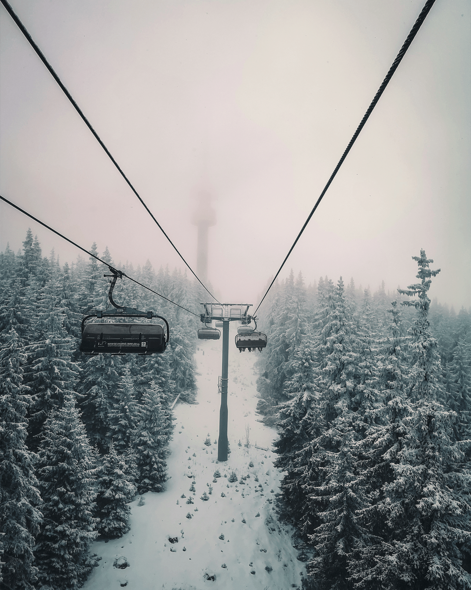 Ski lift ascending through snowy, mist-covered pine forest with fog obscuring distant mountains.