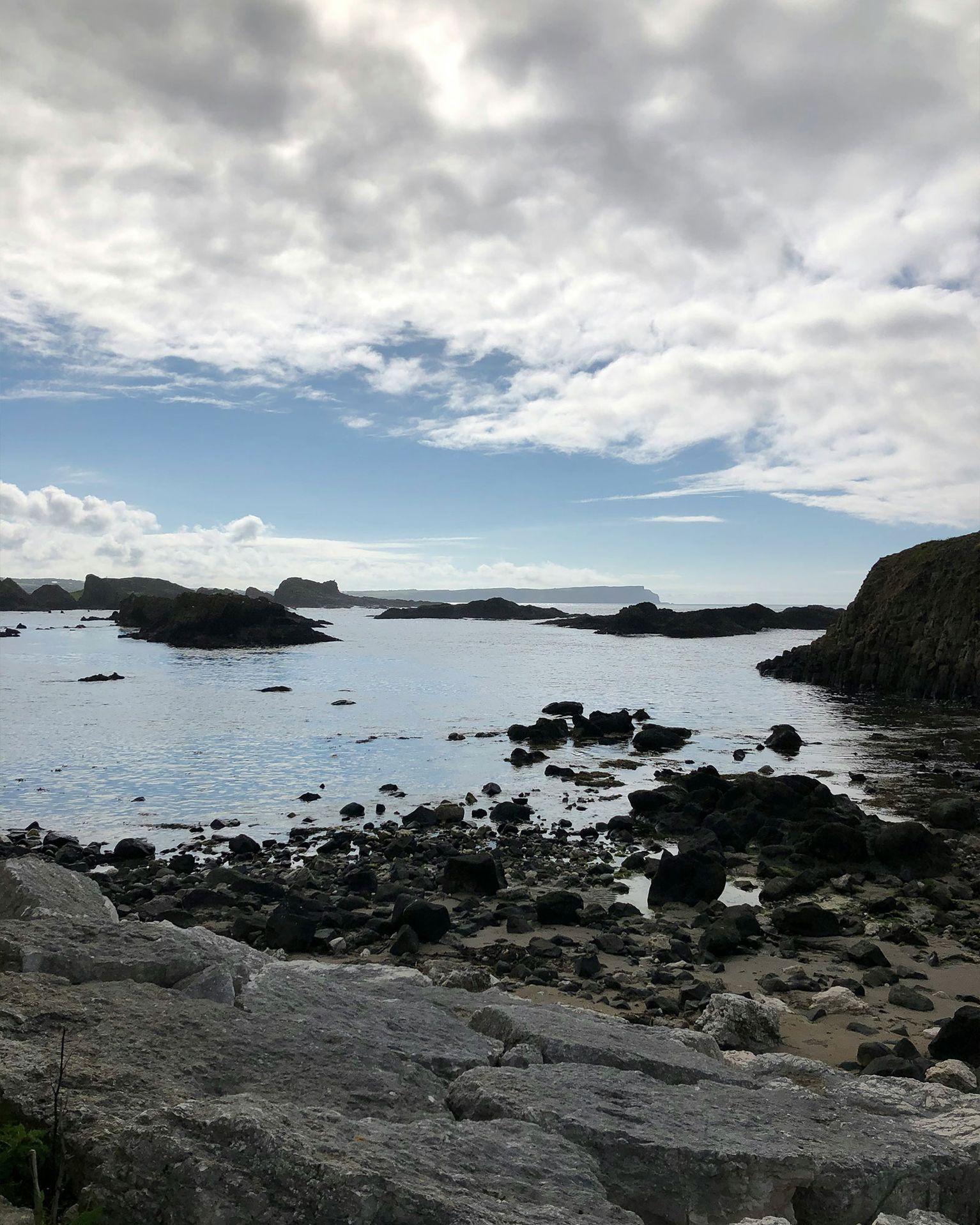 Rocky coastline with calm sea under a cloudy sky, scattered rocks leading to the water's edge and distant land on the horizon.