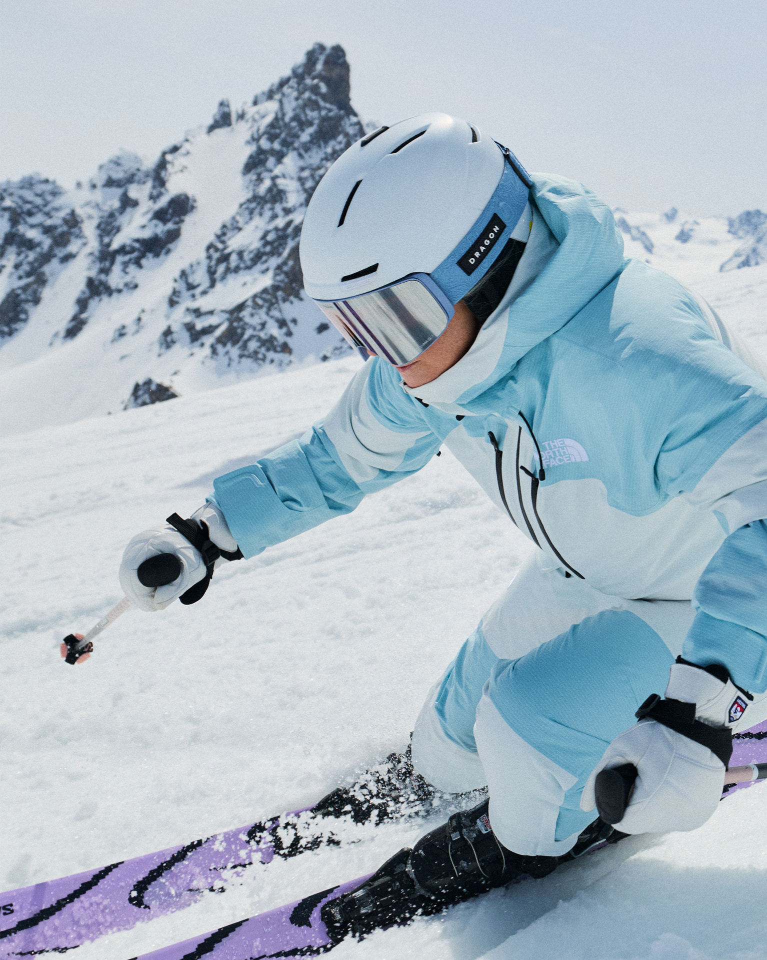 A skier in a blue suit and white helmet skiing down a snowy slope, with rugged mountain peaks in the background.