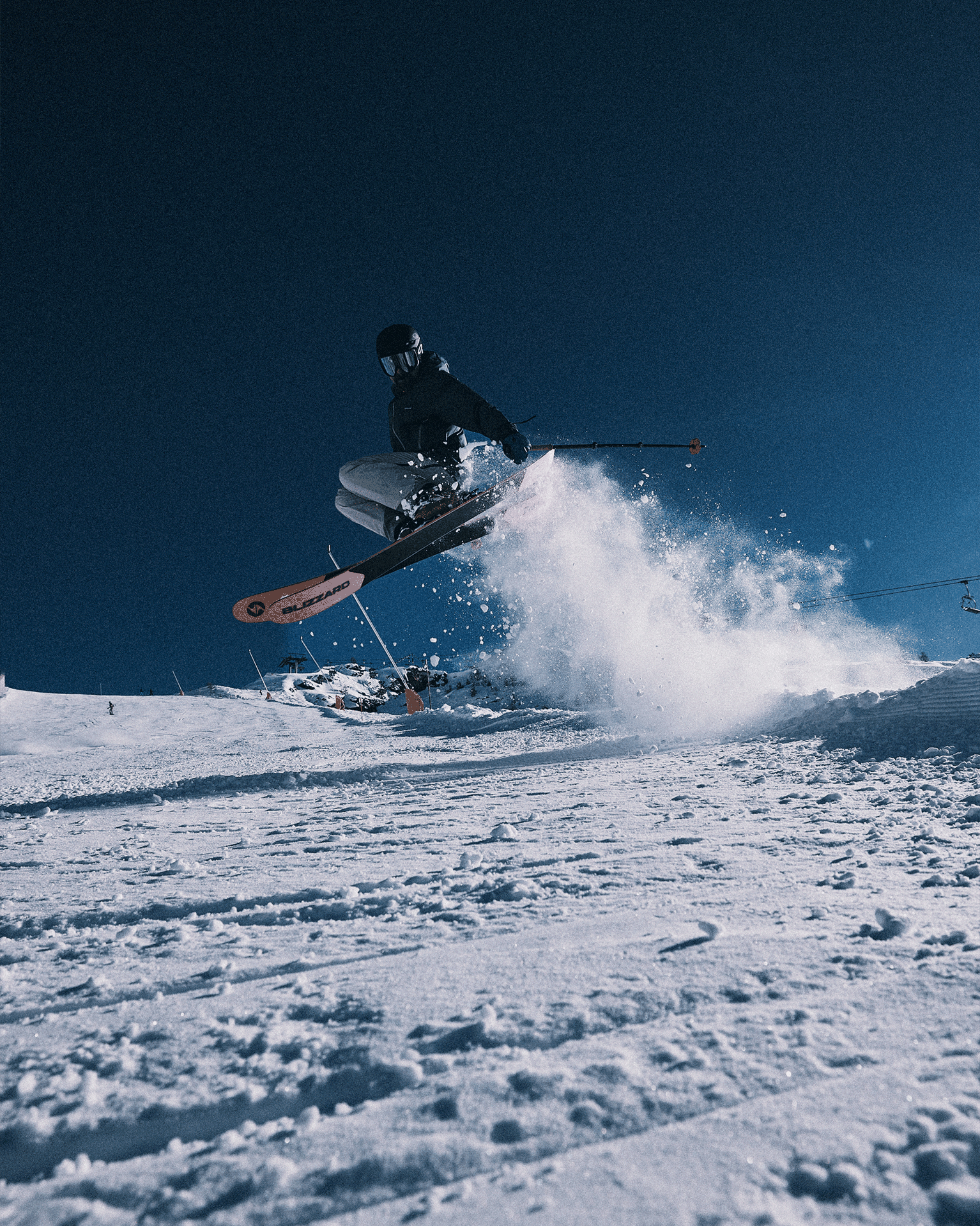 Skier catching air mid-jump on a snowy slope, cloud of powder trailing, under a clear blue sky.