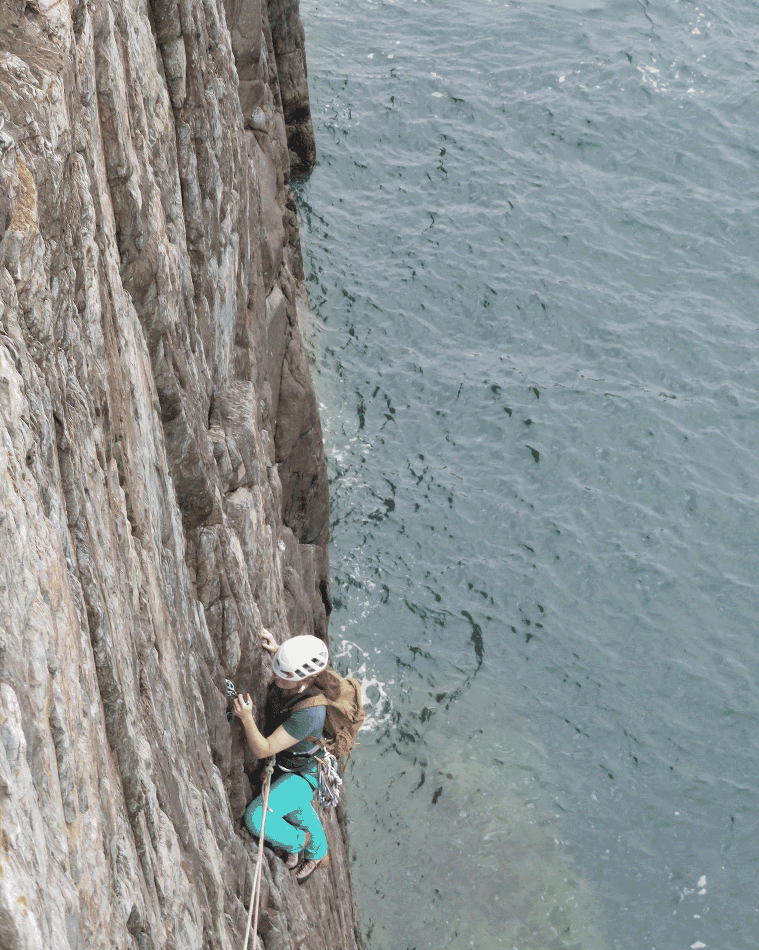 A climber with a helmet and gear scales a steep rock face above water, displaying determination and skill.
