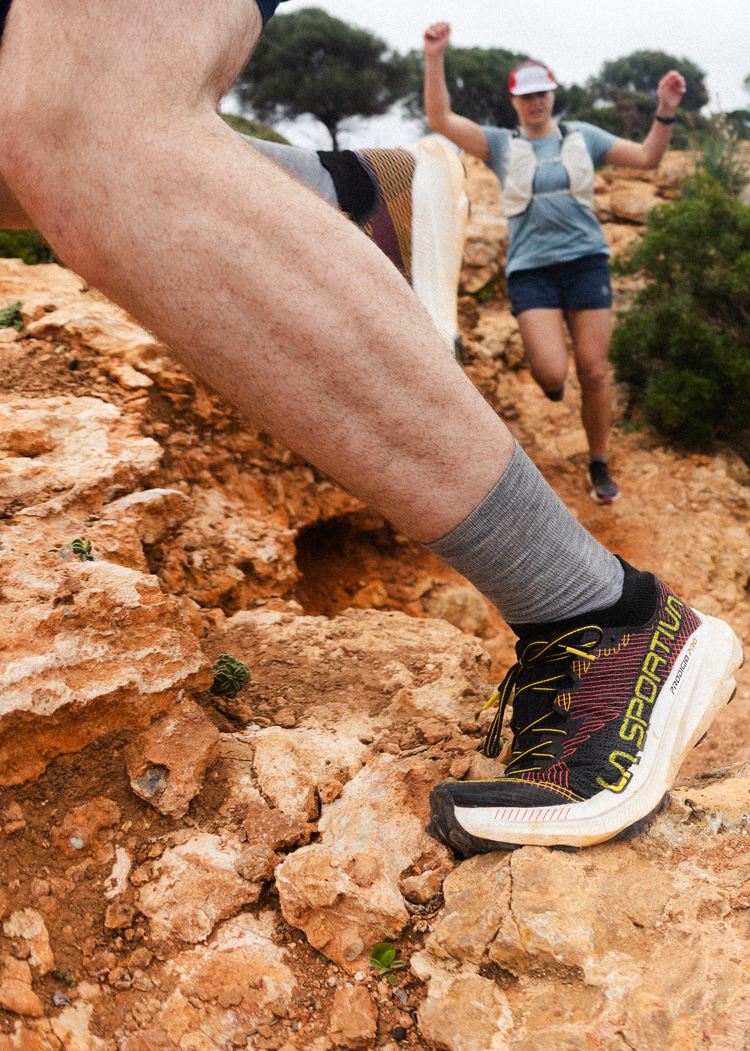Trail runner descending rocky path, focus on La Sportiva shoe. Another runner in the background, wearing athletic gear, cheering energetically.