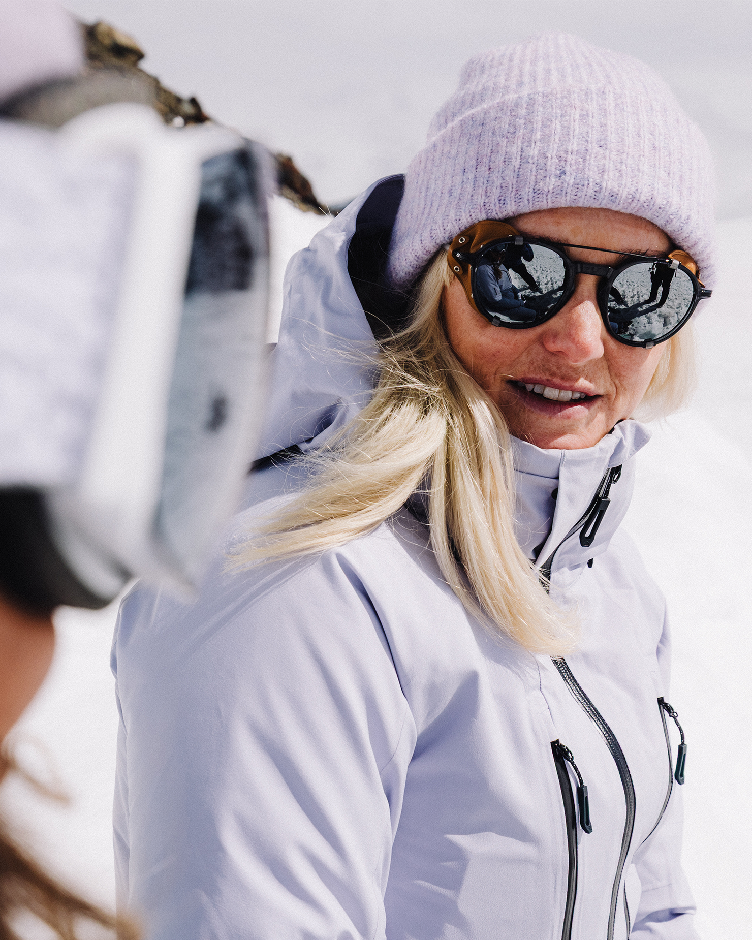 Woman in a light purple jacket and beanie smiling on a snowy mountain, wearing reflective sunglasses.