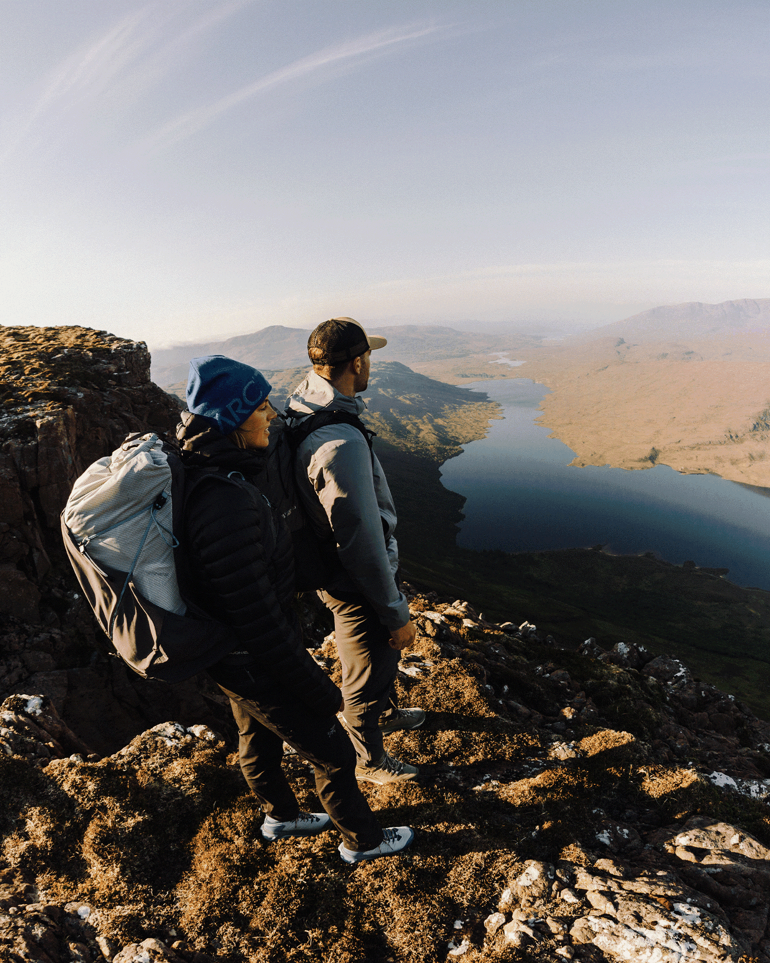 Two hikers in outdoor gear stand on a rocky cliff, overlooking a vast lake and mountainous landscape in clear weather.