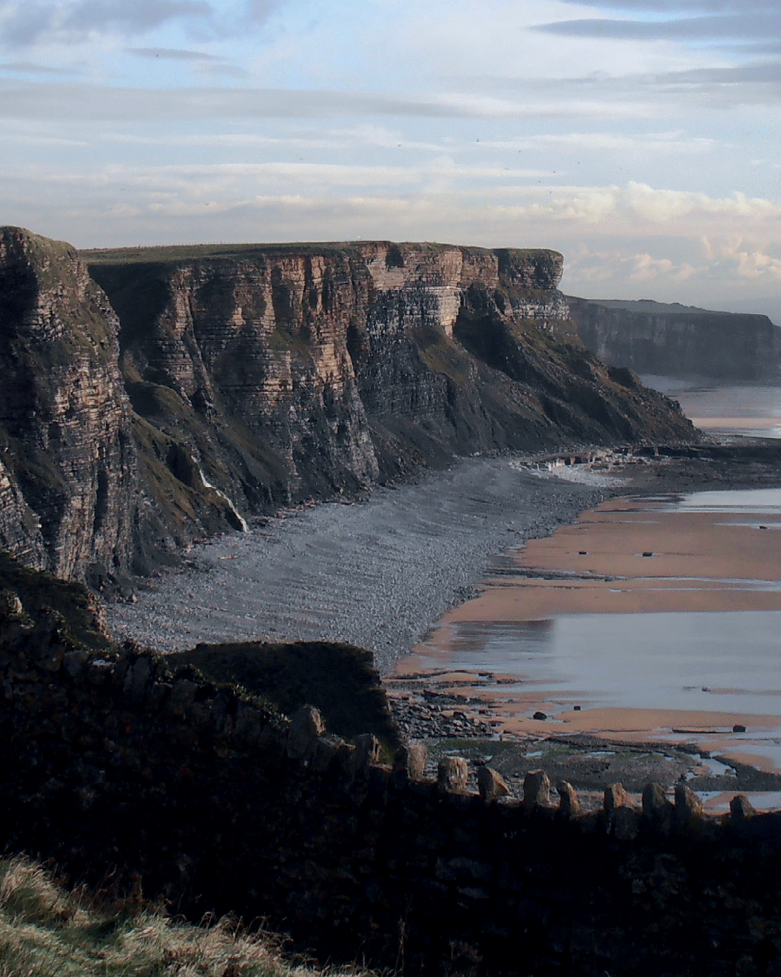 Dramatic coastal cliffs overlooking a pebble beach and sea, with a stone wall in the foreground under a partly cloudy sky.