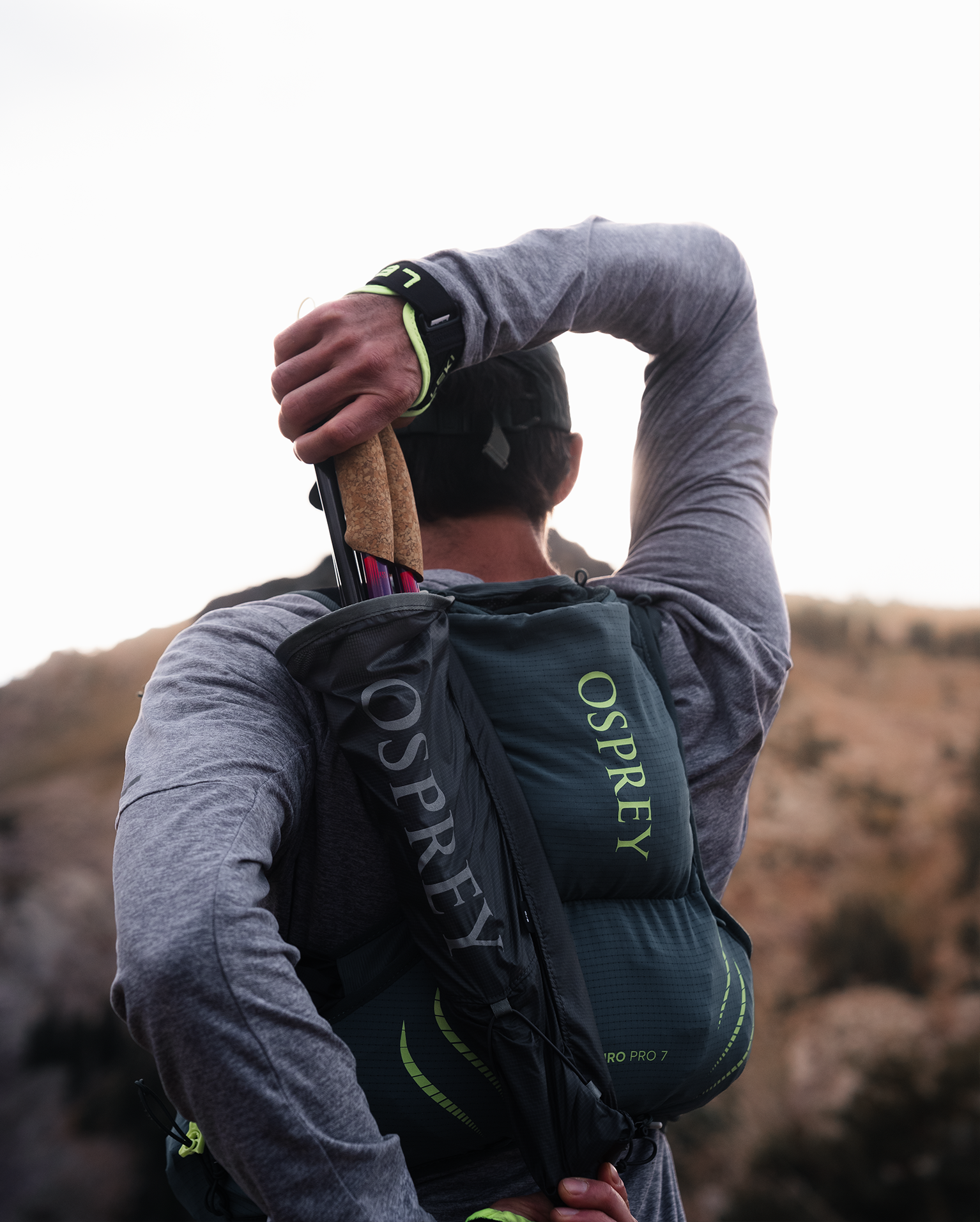Man in grey shirt stretches arm, holding trekking poles and wearing an Osprey backpack against a mountain backdrop.