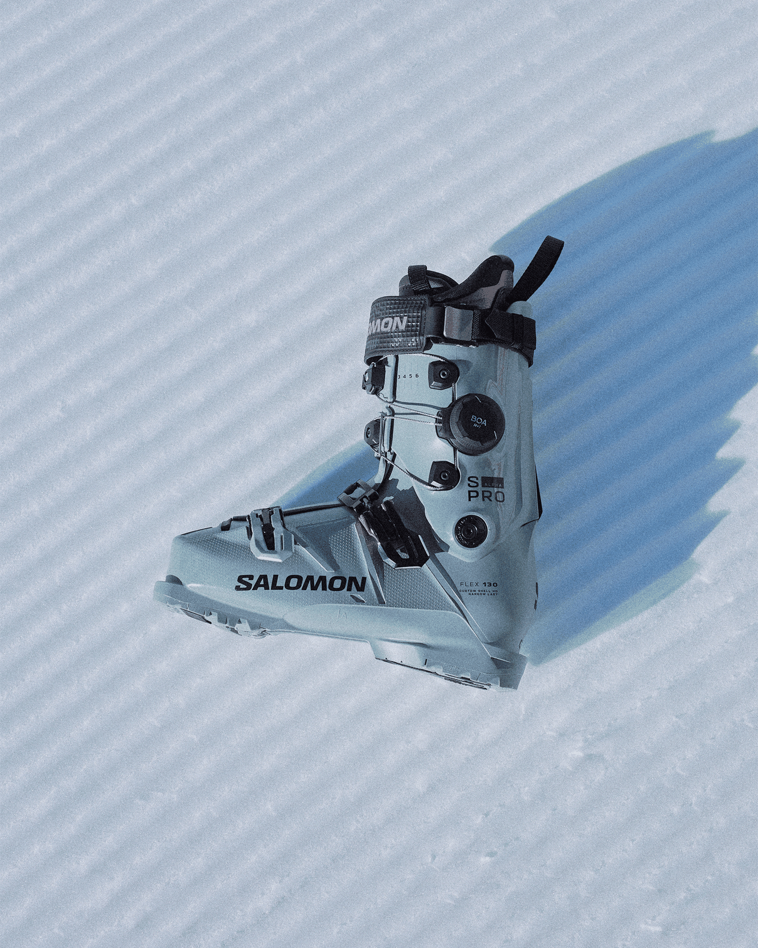 Grey ski boot with black details resting on textured snow, featuring adjustment dials and fastenings, branded with "Salomon S/PRO".