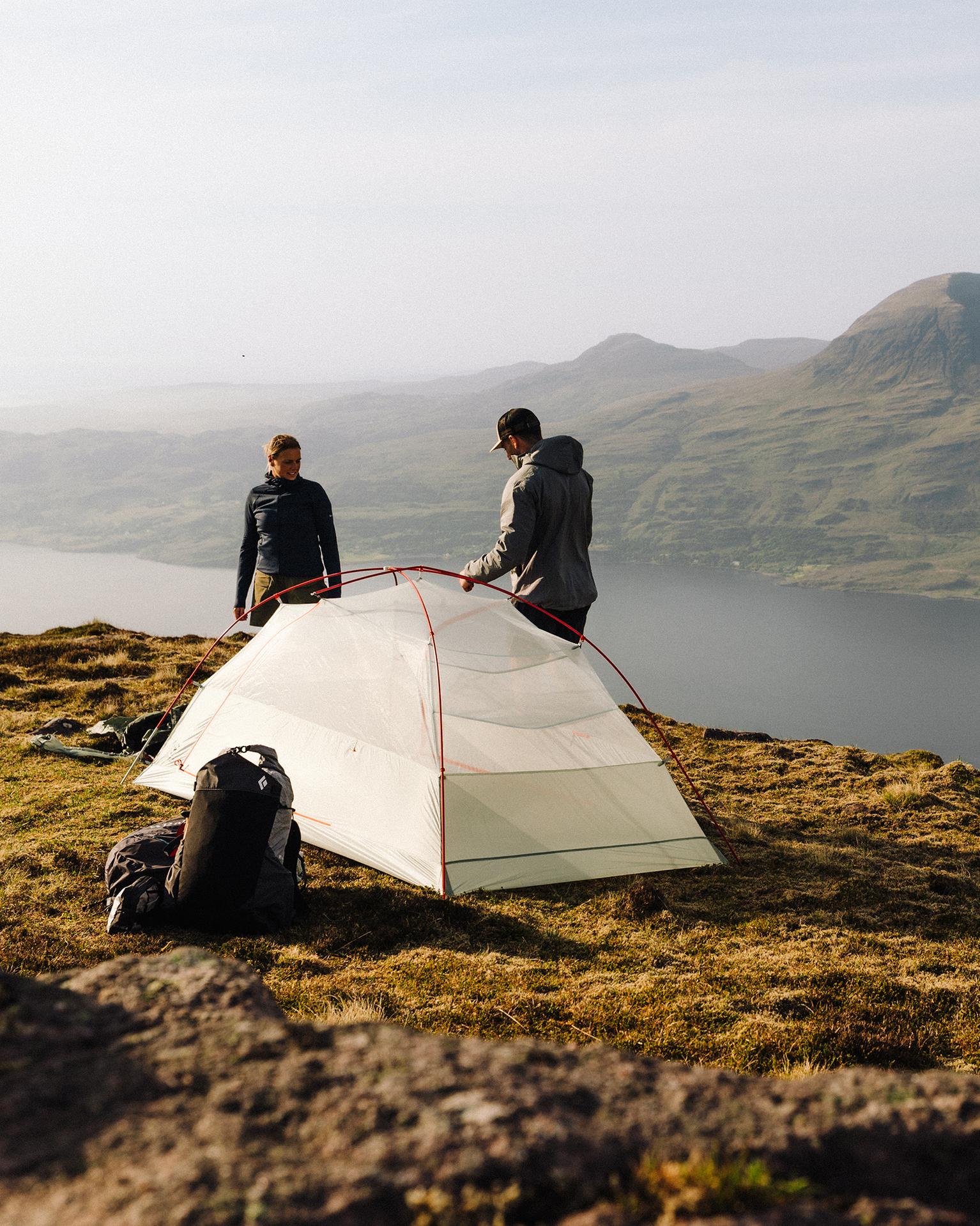 Two people setting up a tent on a grassy hilltop overlooking a scenic lake and distant mountains. A backpack rests nearby.