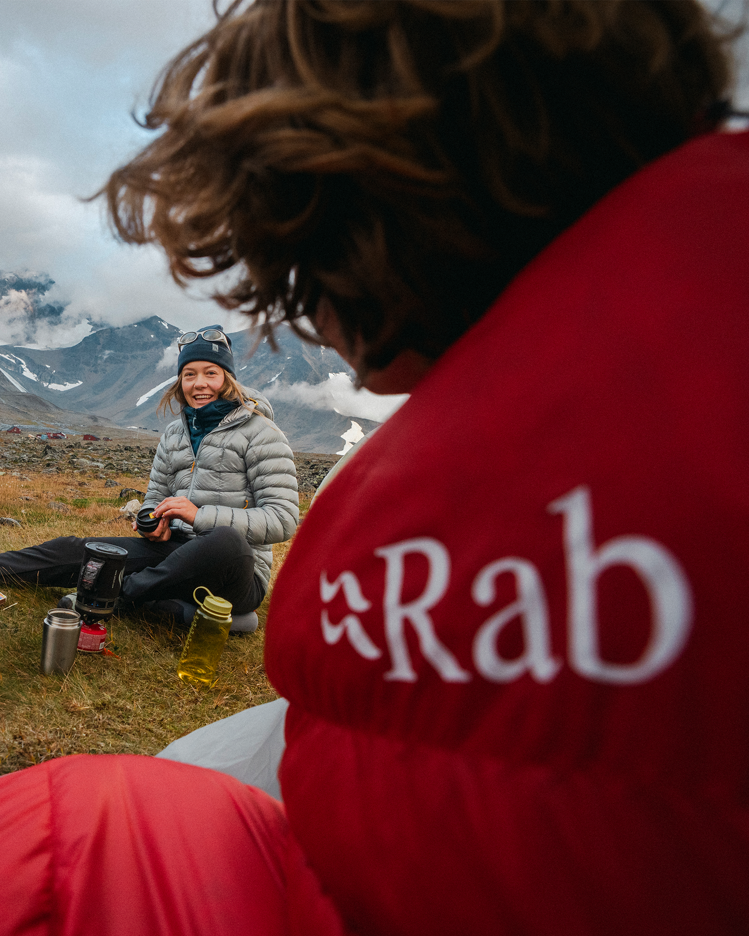 People in warm jackets relax outdoors in a mountainous landscape, with camping gear and cloudy skies in the background.