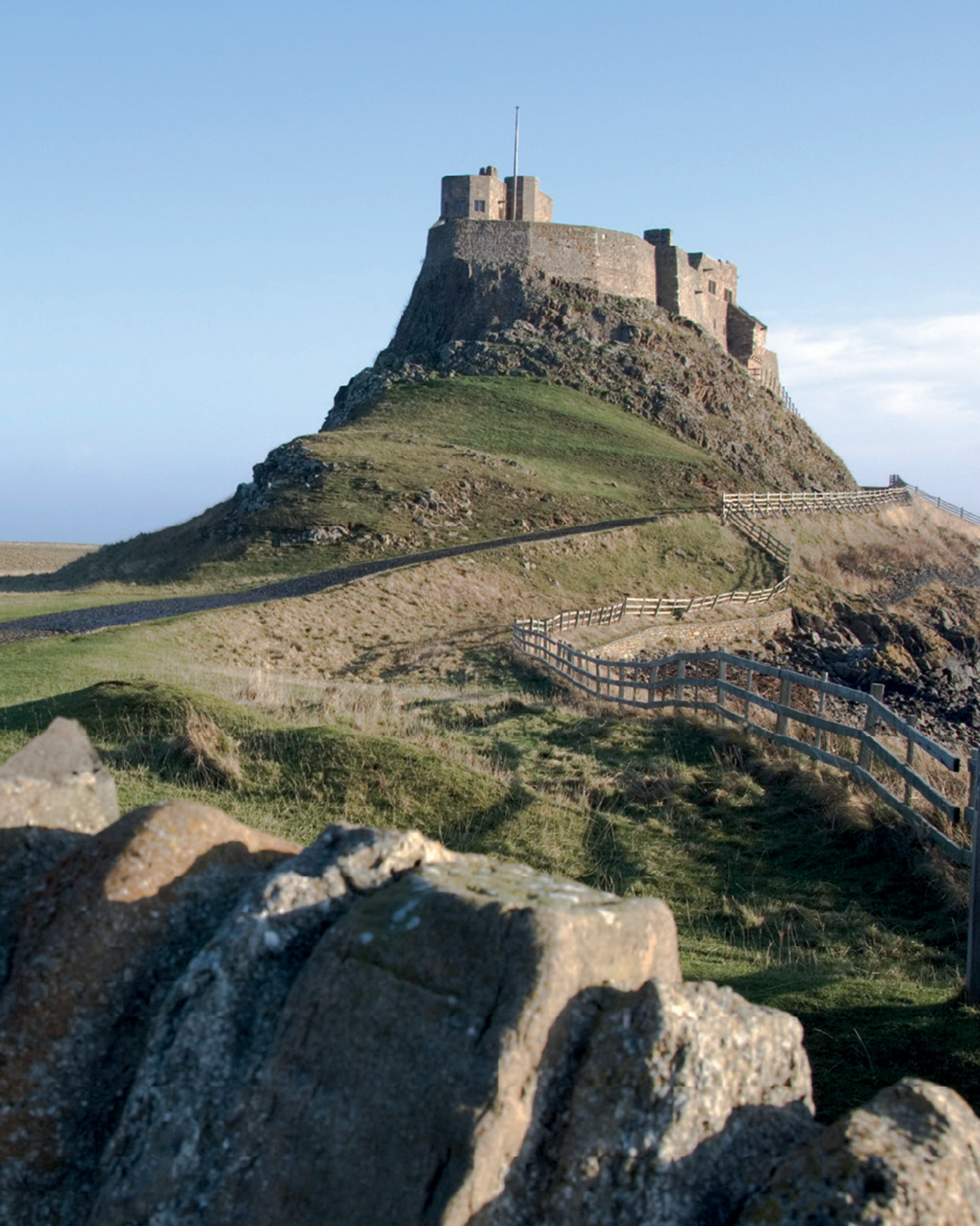 A historic castle perched on a steep hill, with a winding path and wooden fence leading up to it under a clear blue sky.
