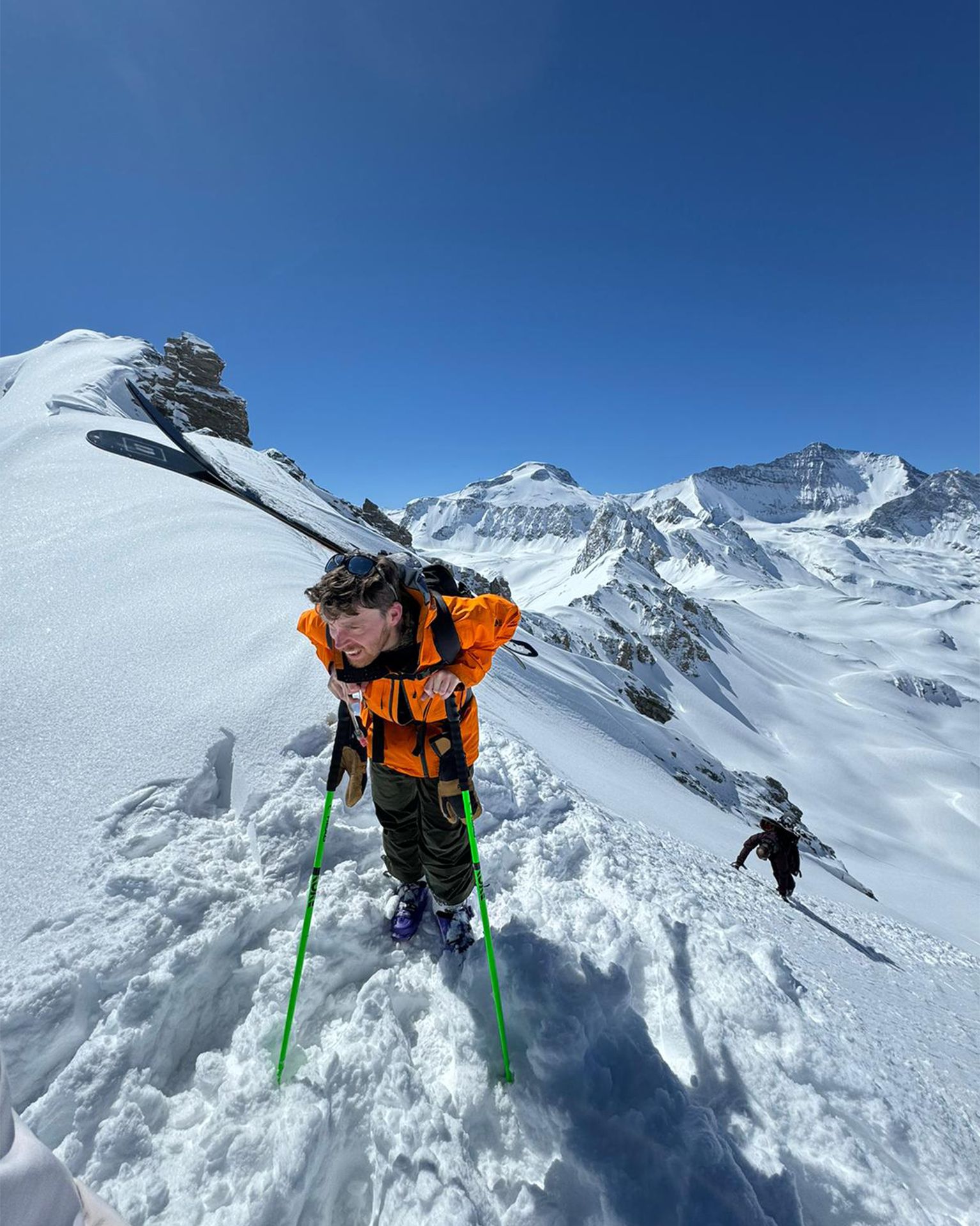 Person in bright orange jacket climbing snowy mountain with green poles under clear blue sky. Snow-covered peaks in background.