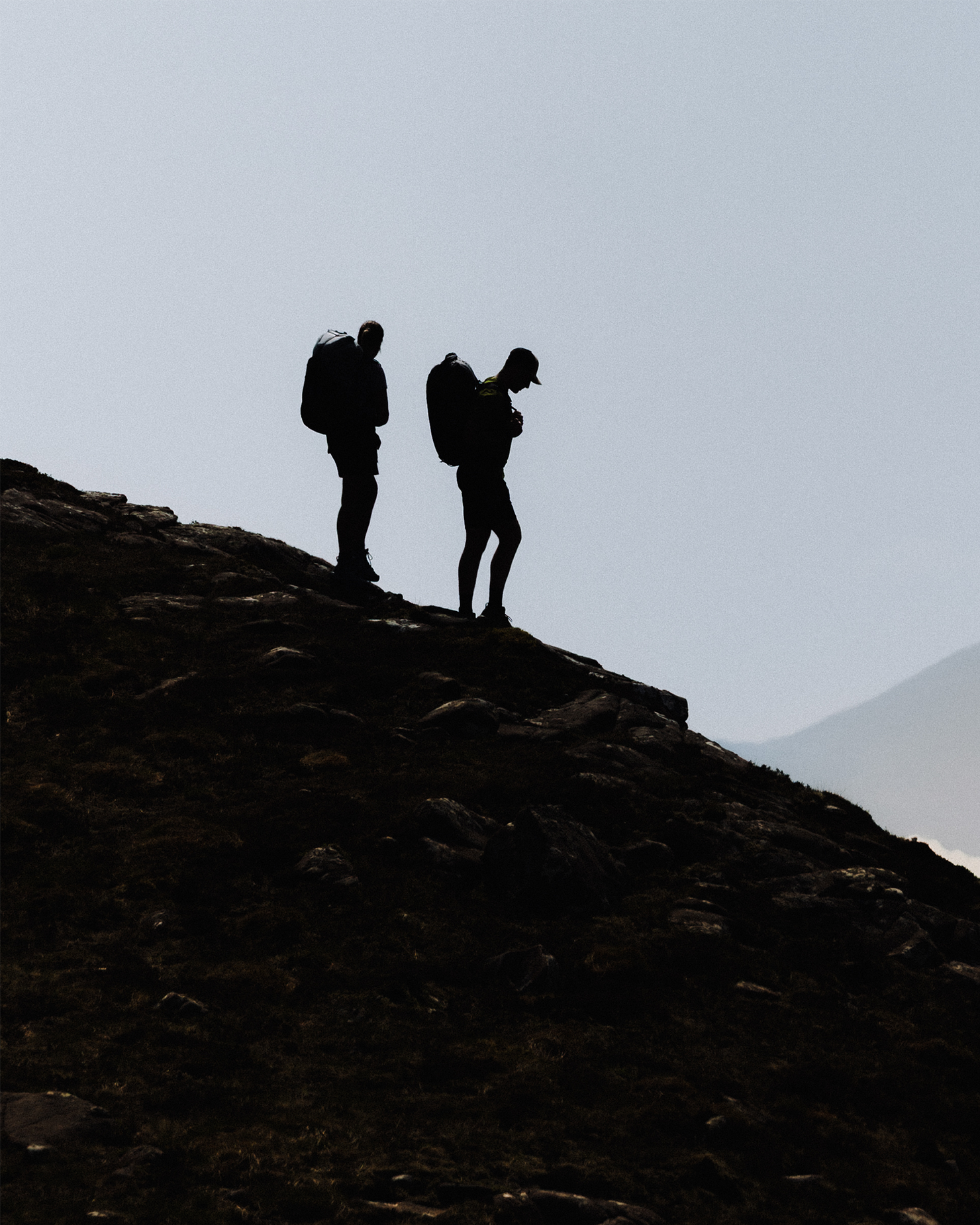 Two silhouetted hikers with backpacks stand on a rocky hillside, captured against a hazy sky.