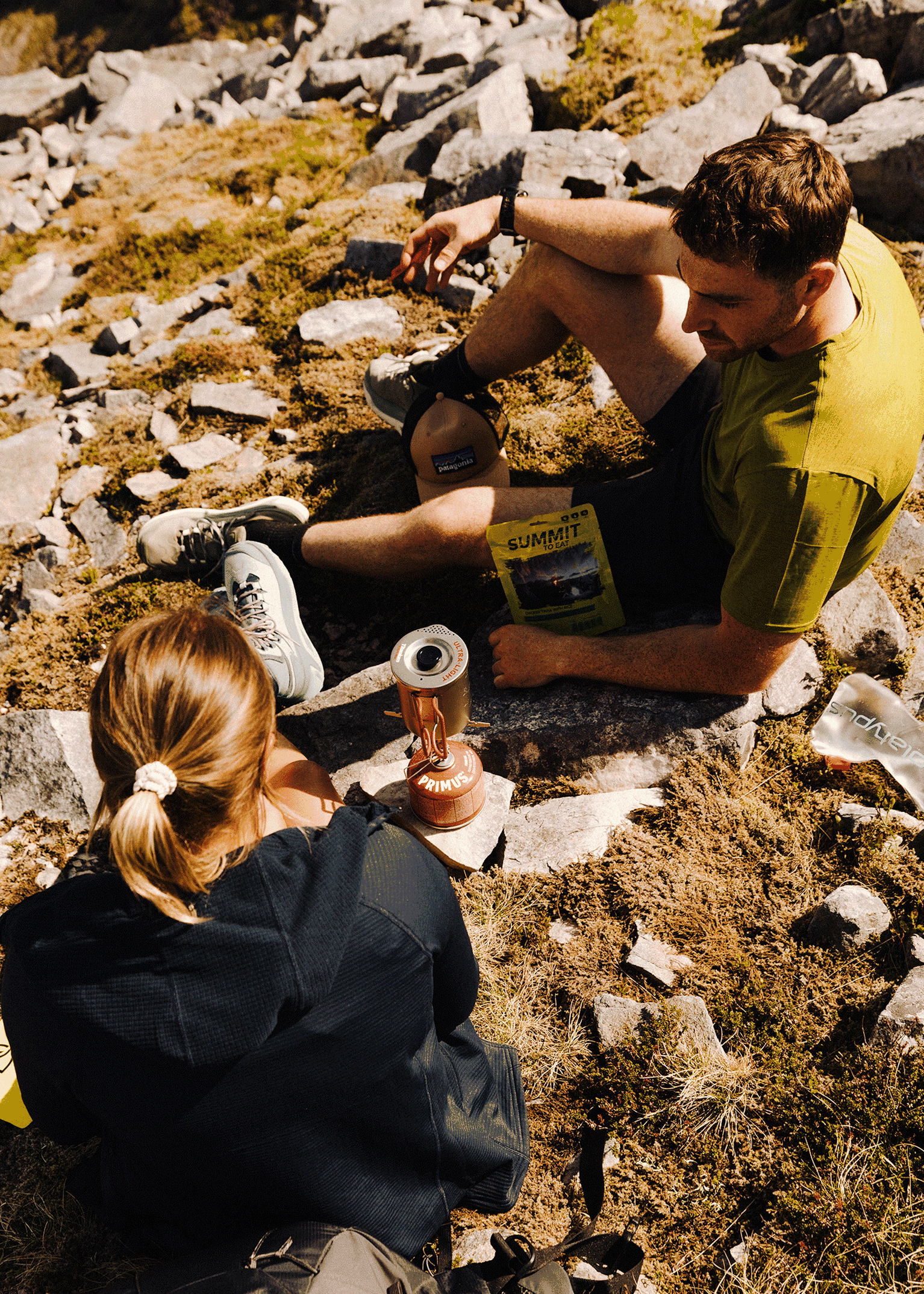 Two hikers rest on rocky terrain, enjoying a trail meal. A portable stove and packaged food are visible beside them.