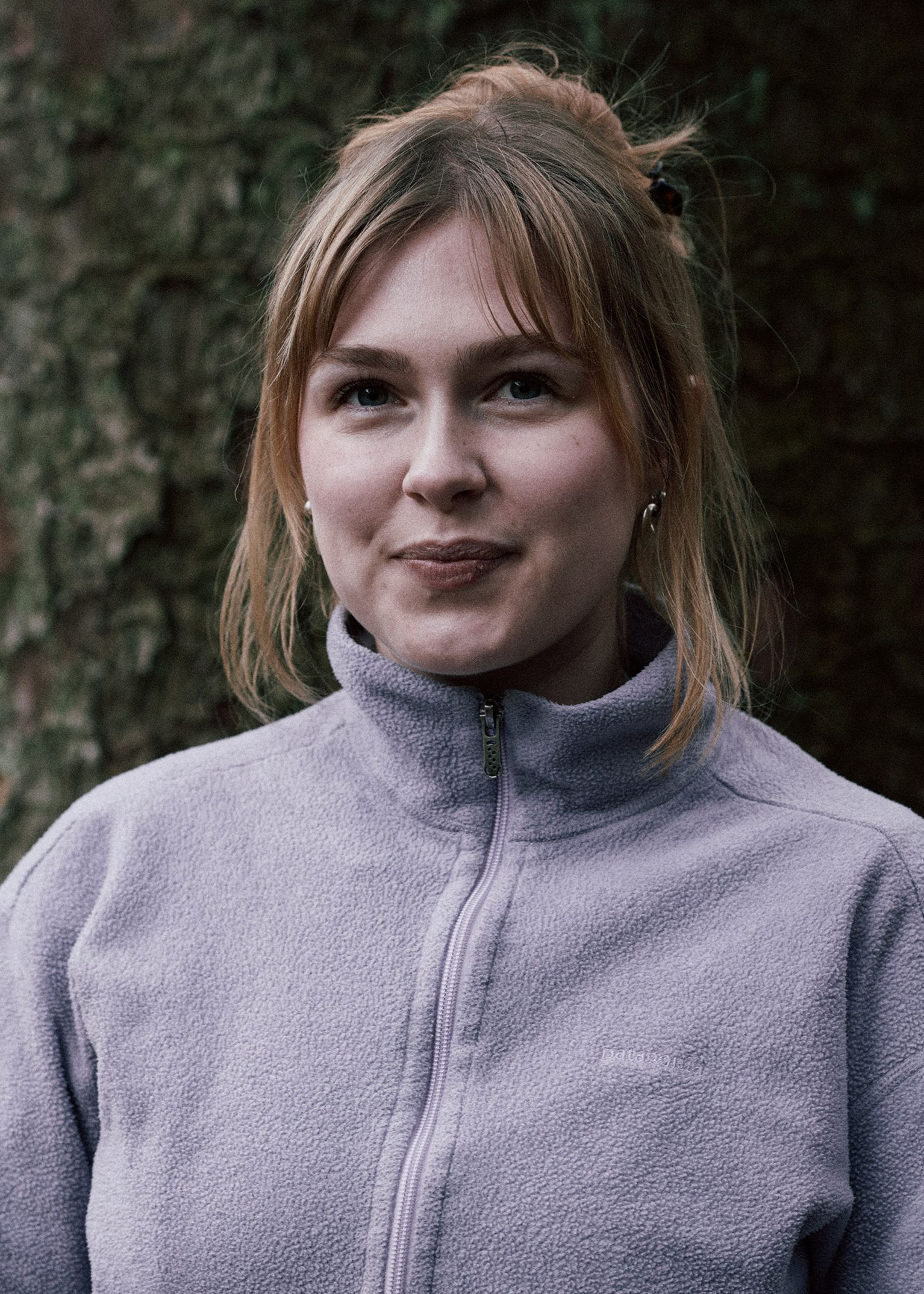 Woman in a light purple fleece jacket stands in front of a tree, smiling softly with her hair tied up.