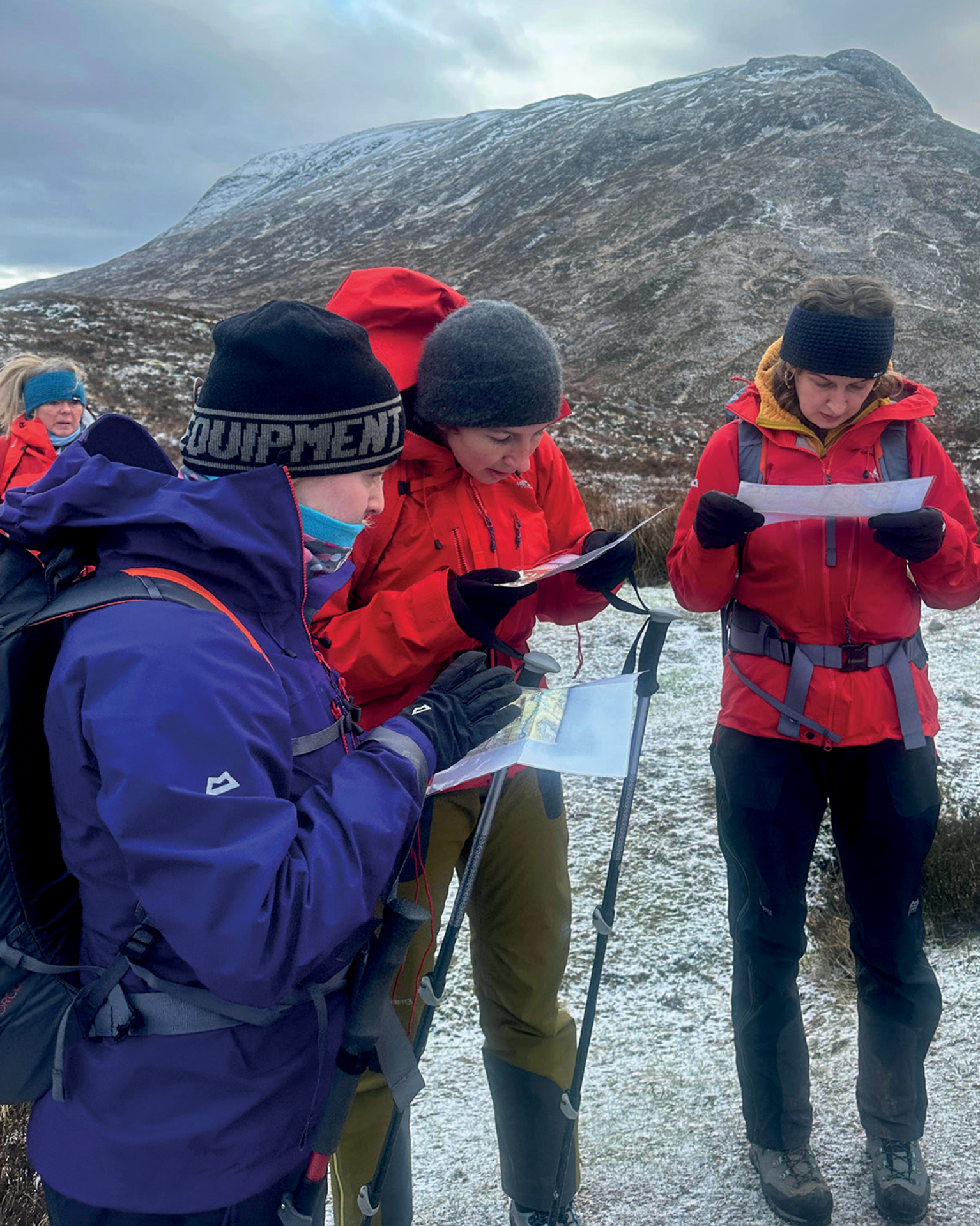 Hikers in winter clothing study maps on a snowy mountain trail, with rugged terrain in the background.