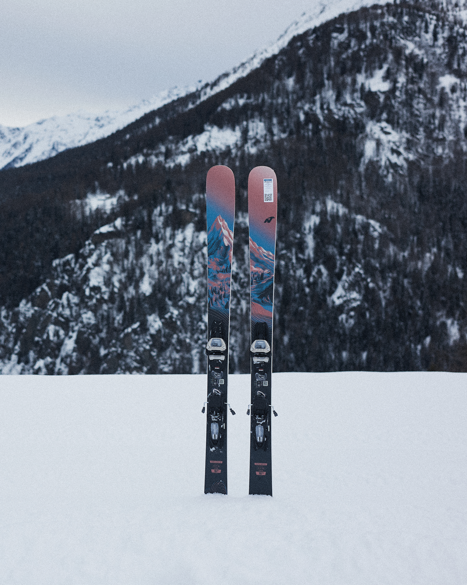 Two colourful skis stand upright in the snow against a backdrop of snow-covered mountains.