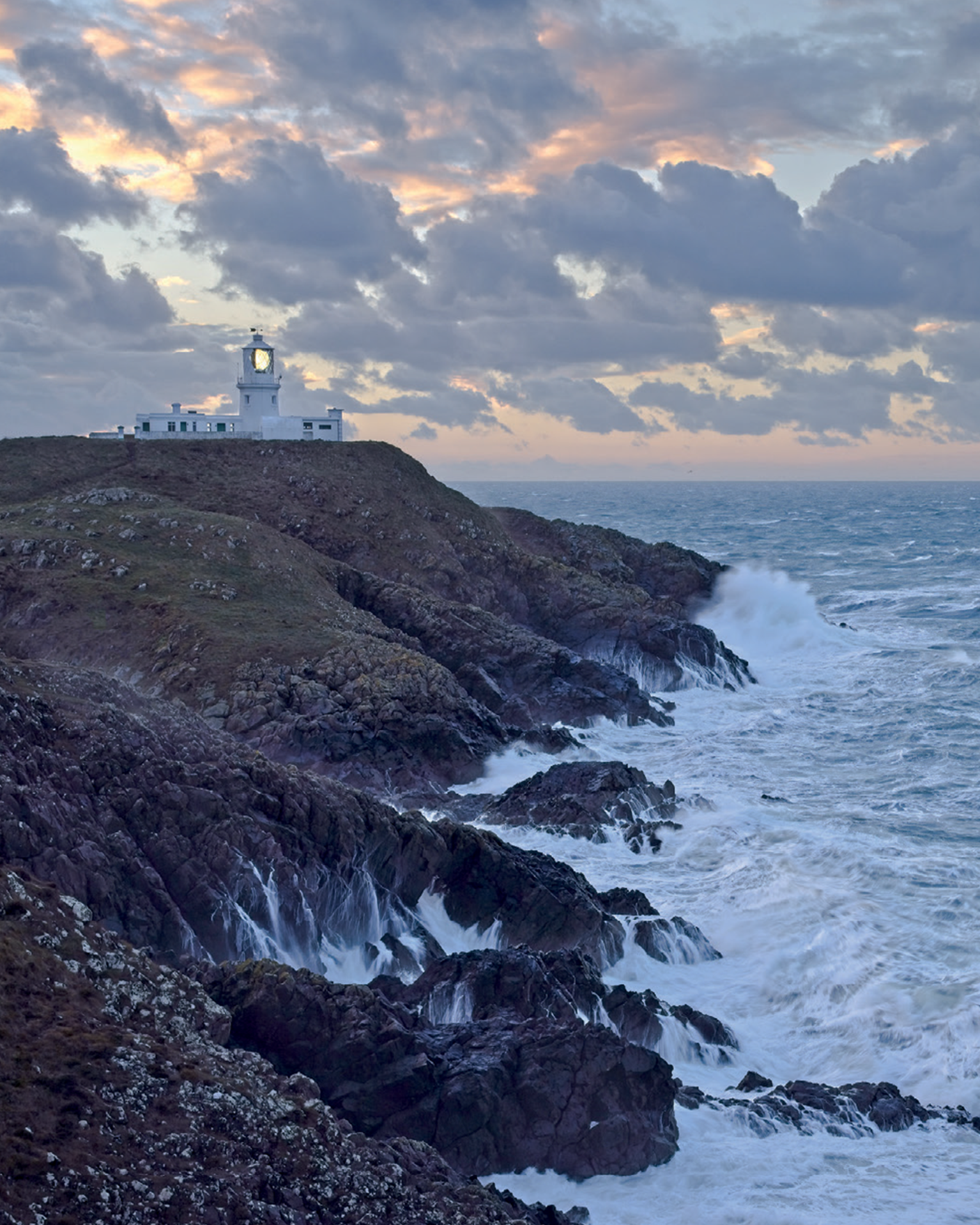 Dramatic coastal scene with waves crashing against rocky cliffs, a white lighthouse stands atop the hill under a cloudy sunset sky.