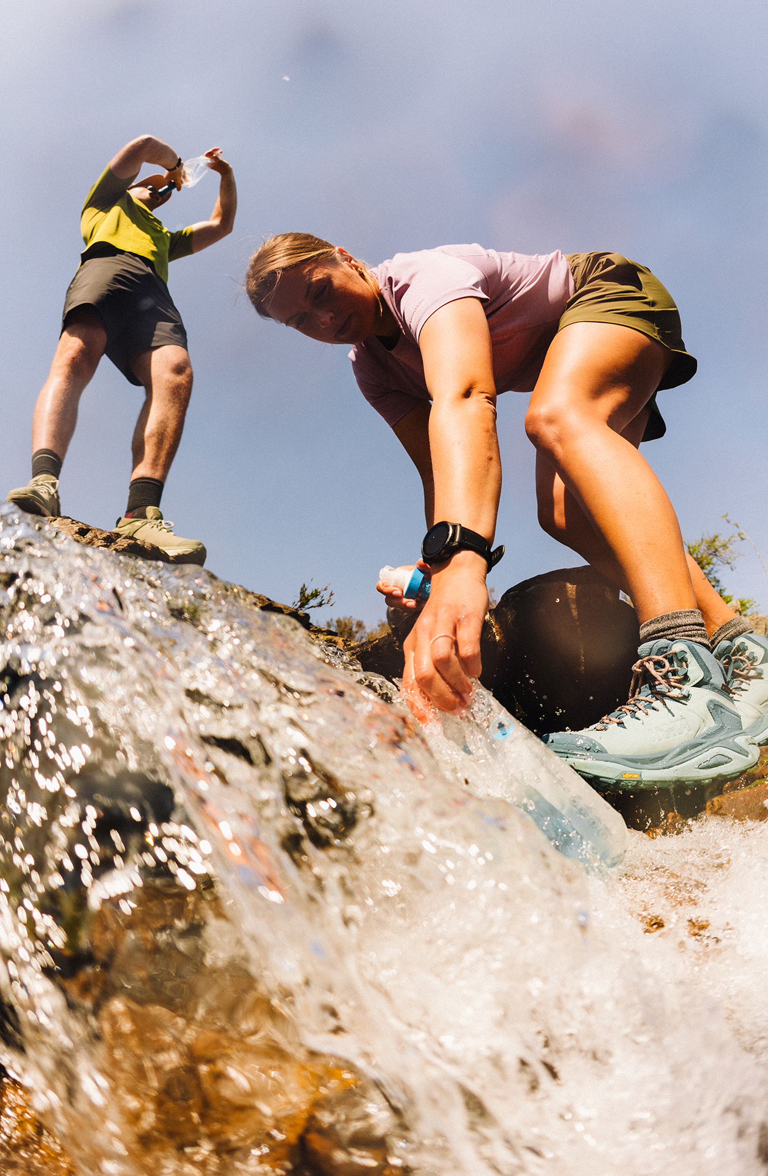 Two people collect water from a flowing stream. One crouches to fill a bottle, while the other stands nearby, holding a camera.