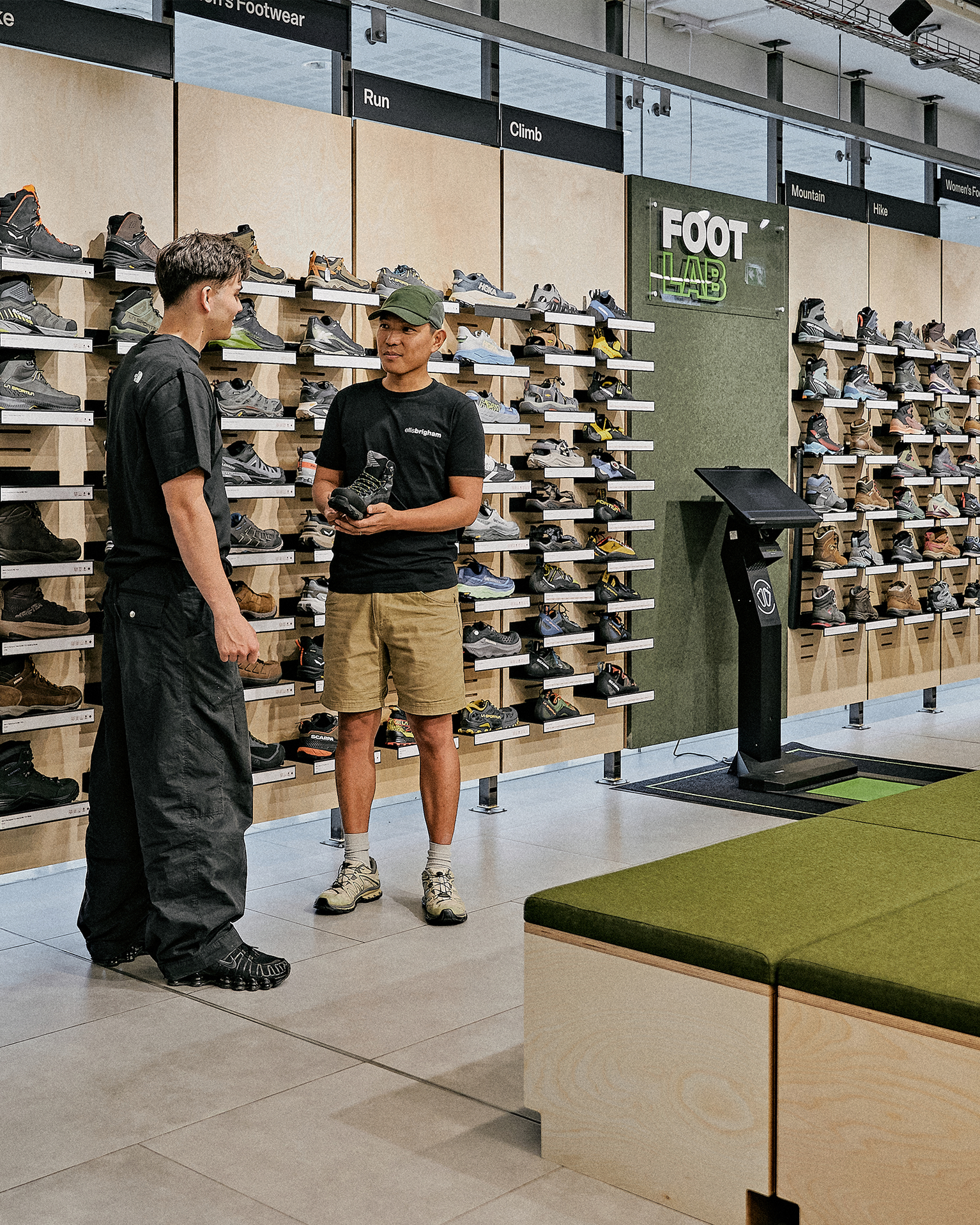 Two men discussing footwear in a mountain sports shop, surrounded by shelves of shoes and an interactive kiosk.