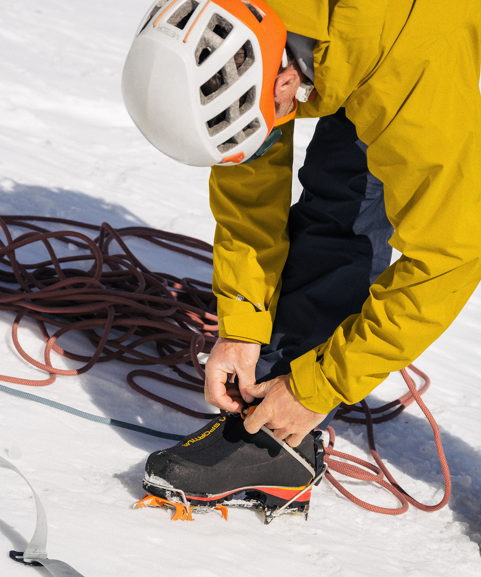 A person in a yellow jacket and helmet adjusts crampons on their boot, surrounded by climbing ropes on snowy terrain.