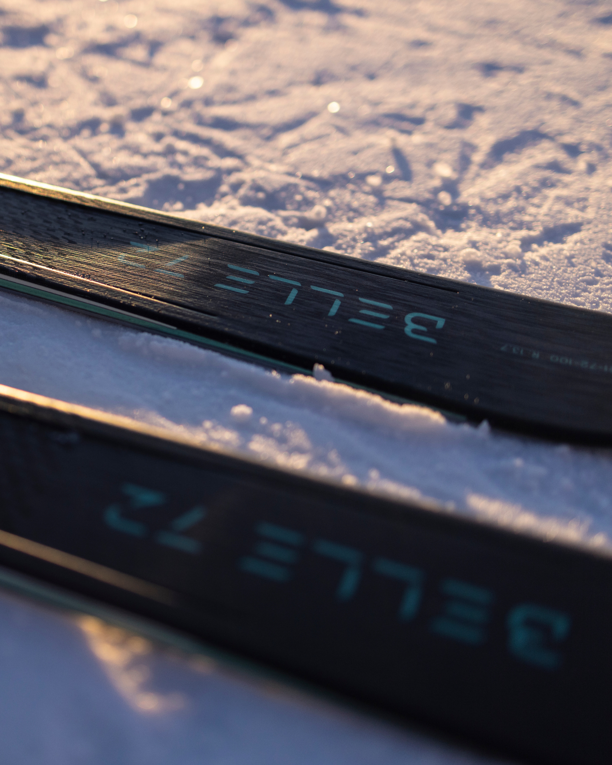 Close-up of ski tips on snow, with "BELLE 72" visible. Sunlight highlights snow texture, creating a crisp, wintery scene.