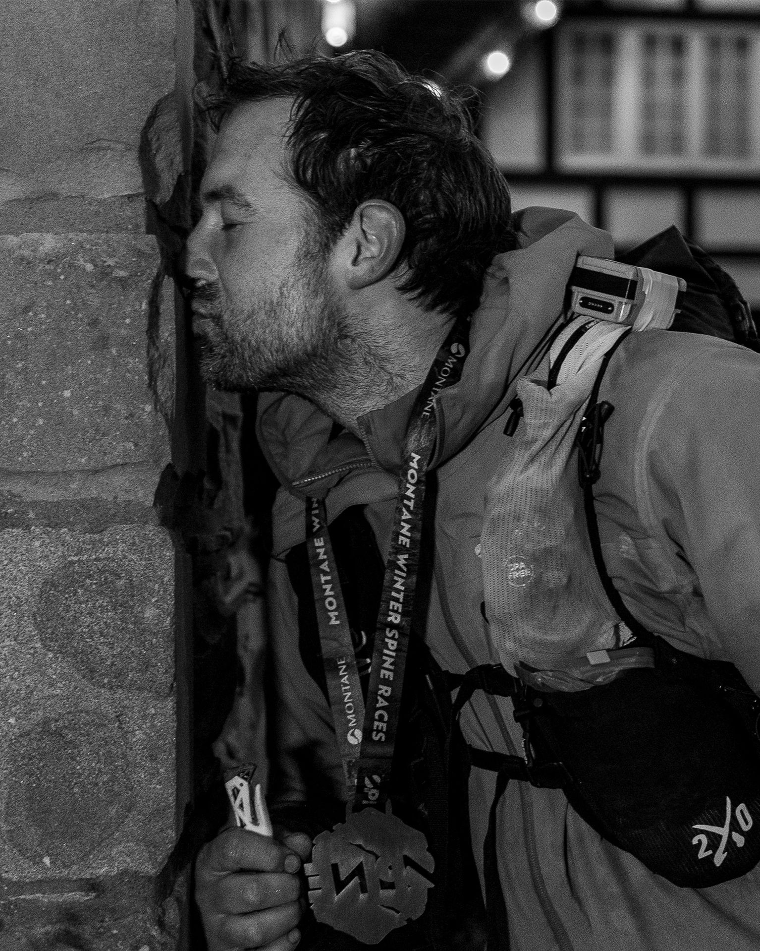 Man in mountain gear, eyes closed, touching forehead to brick wall, wearing medals; appears relieved and contemplative after a race.