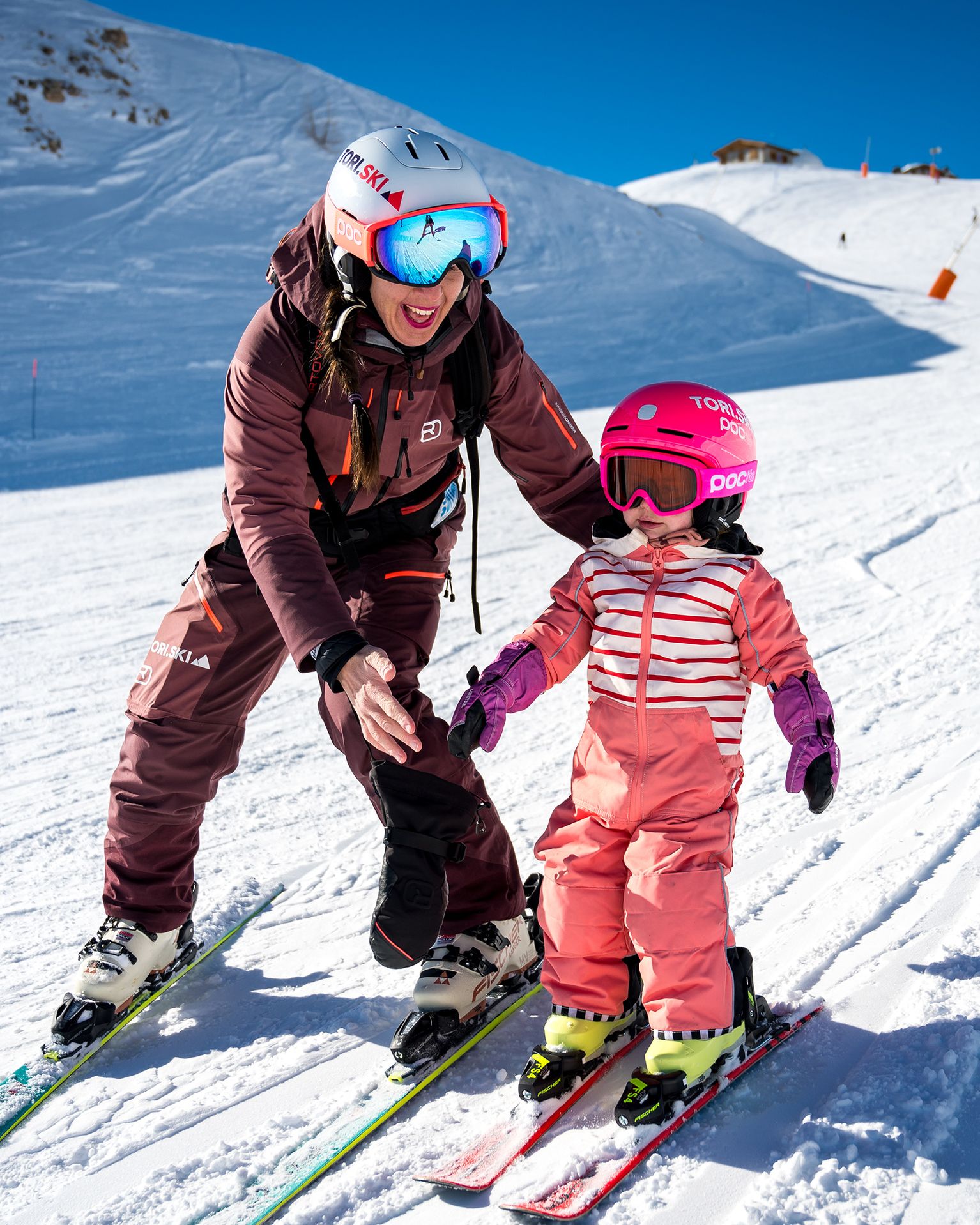 Adult and child skiing on a snowy slope, both wearing helmets and ski gear, with the adult guiding the child.