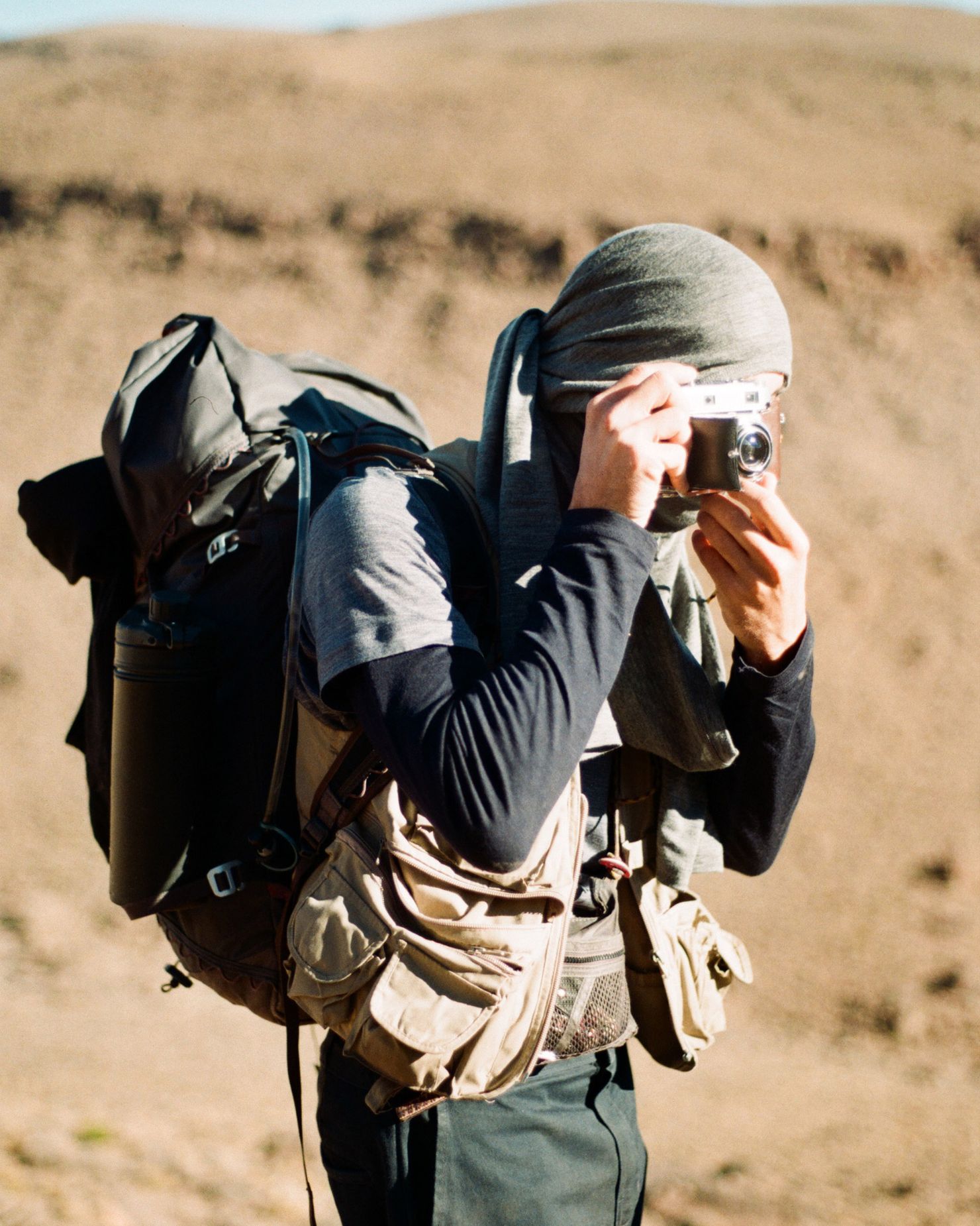 Person with a backpack and headscarf taking a photo in a desert landscape.