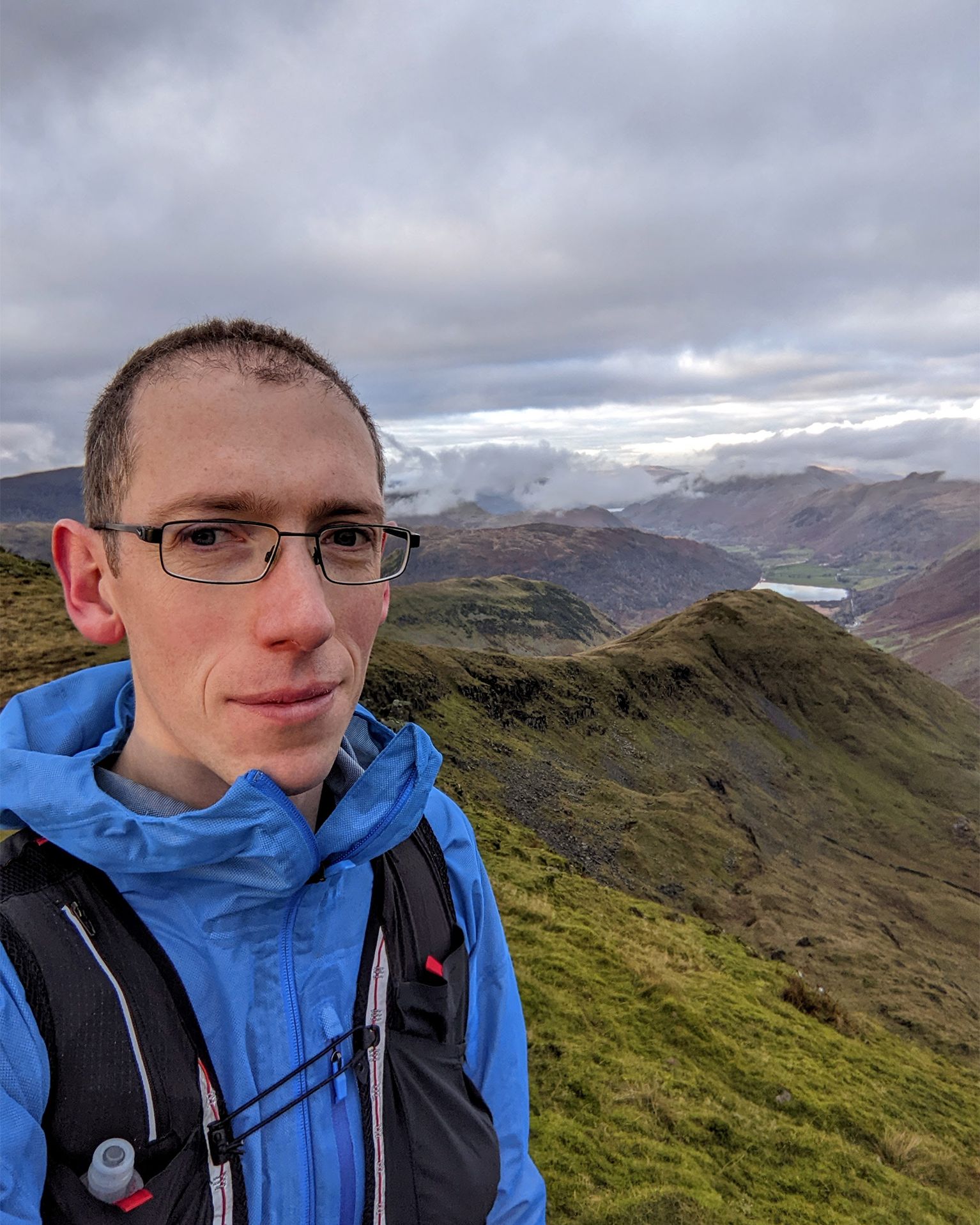 Man in blue jacket and glasses stands on a grassy mountain ridge, with misty valleys and hills in the background. Cloudy sky overhead.