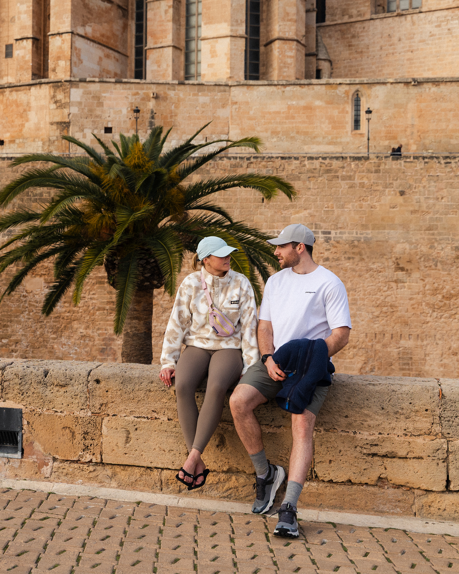 A couple wearing casual clothing and baseball caps sit on a stone wall, chatting, with a historic building and palm tree in the background.