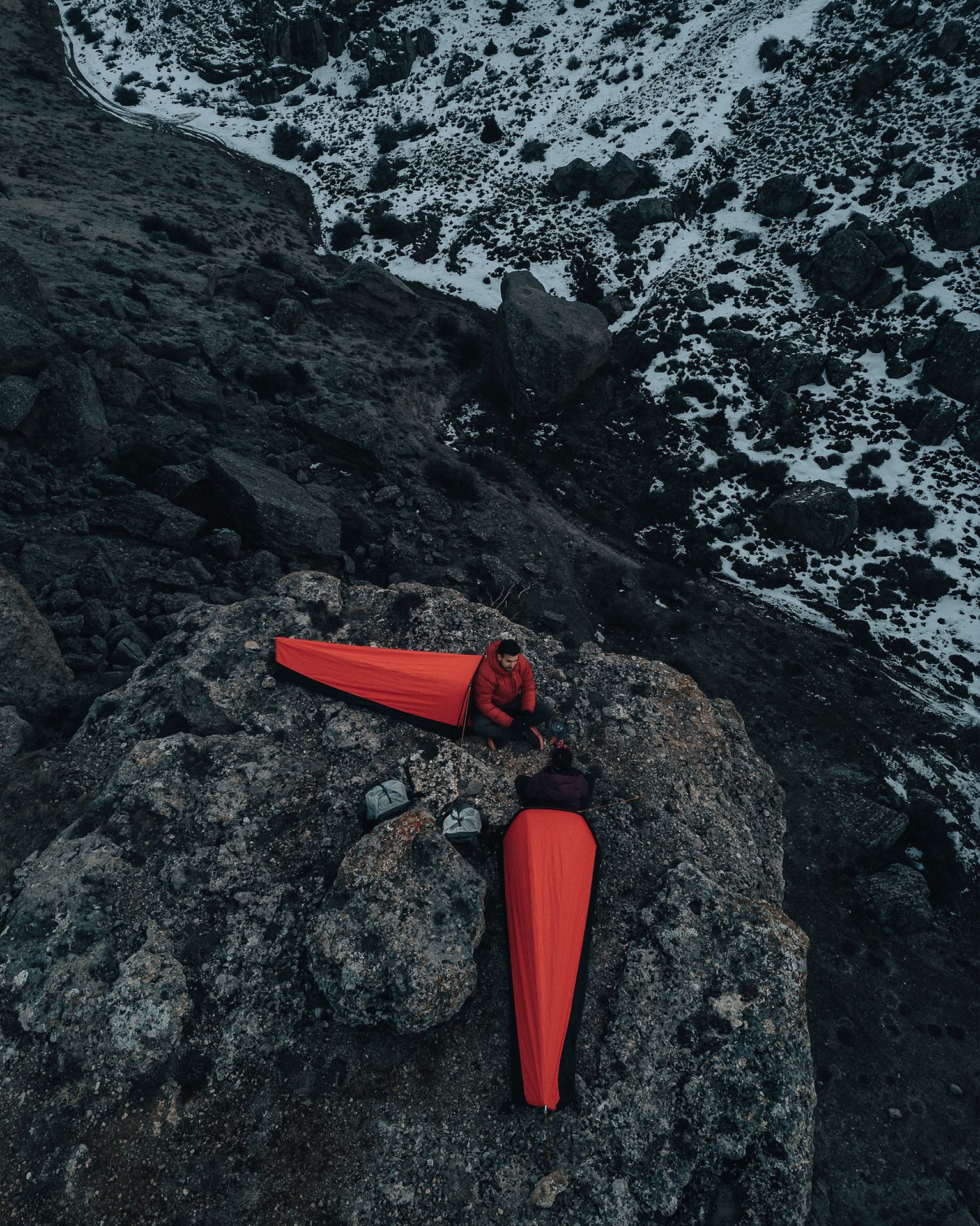 Two people in red bivvy bags camp on a rocky mountain ledge with snow-dusted terrain in the background.