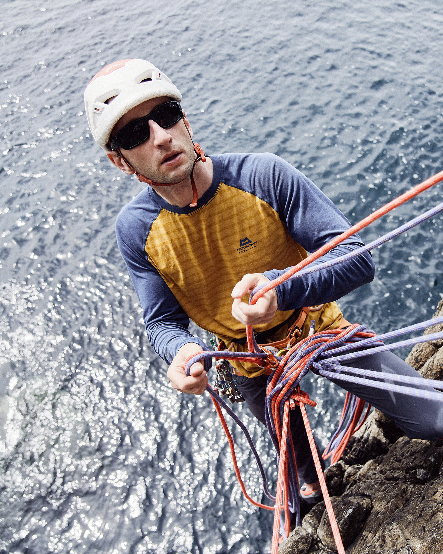 Mountain climber in a yellow and blue top rappelling down a rock face above the ocean, handling ropes with focused determination.